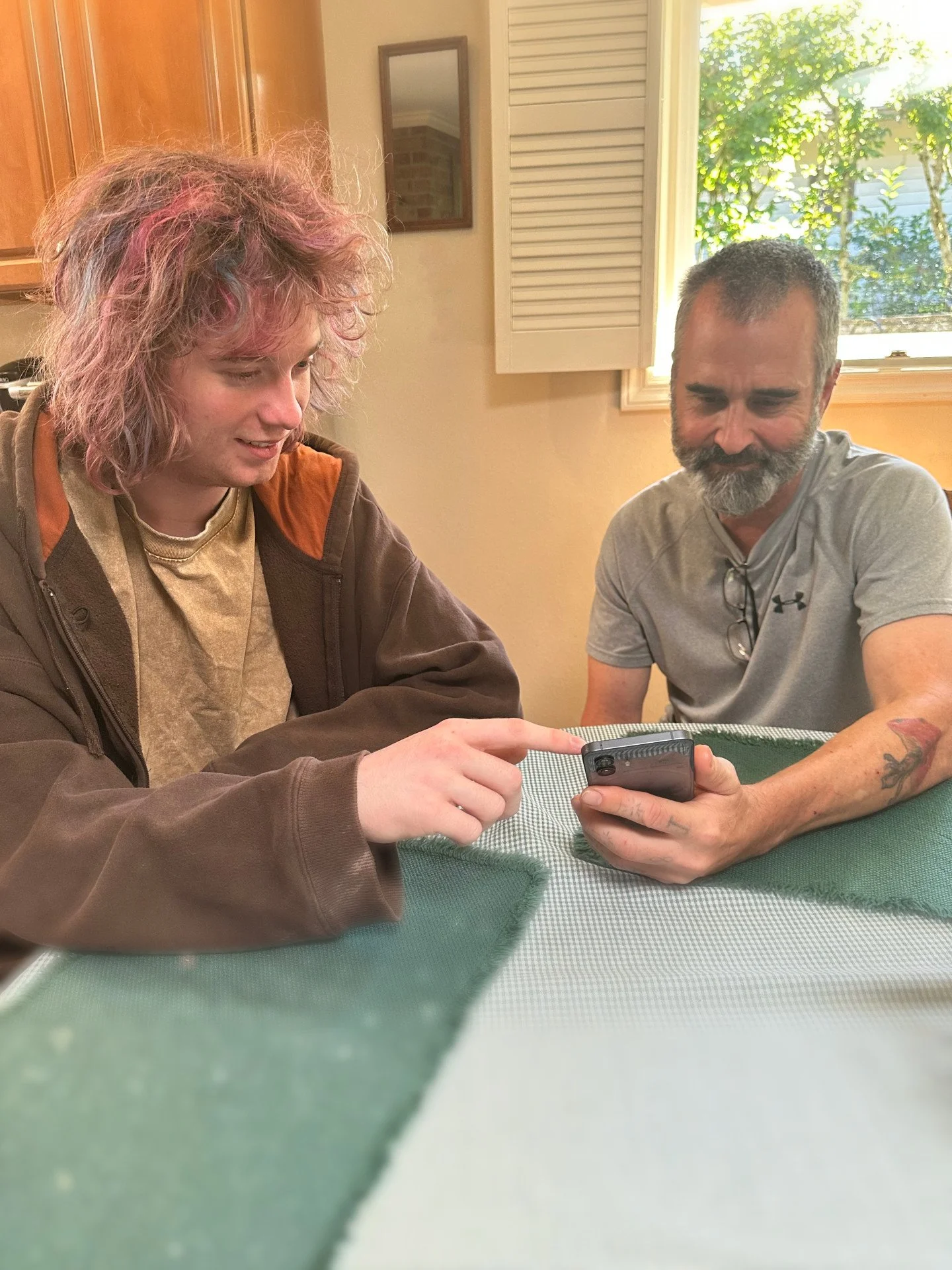 Two people sitting at a table, with Skip showing something to the other while pointing at the phone.