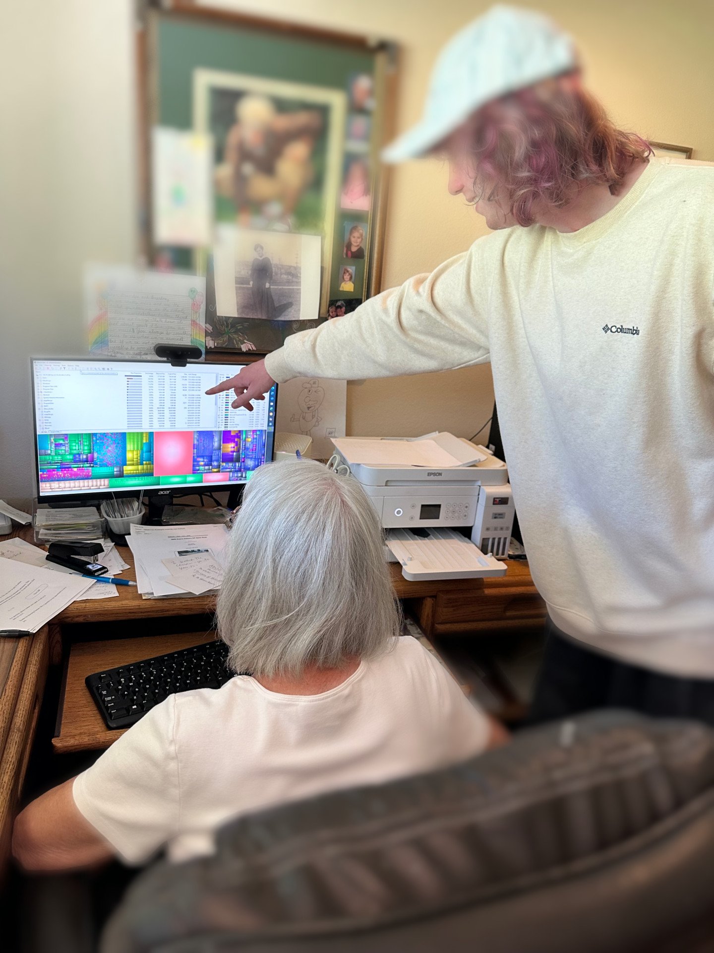 A woman with short gray hair sitting at a desk, looking at a computer screen with a colorful digital file, while Skip points something out.