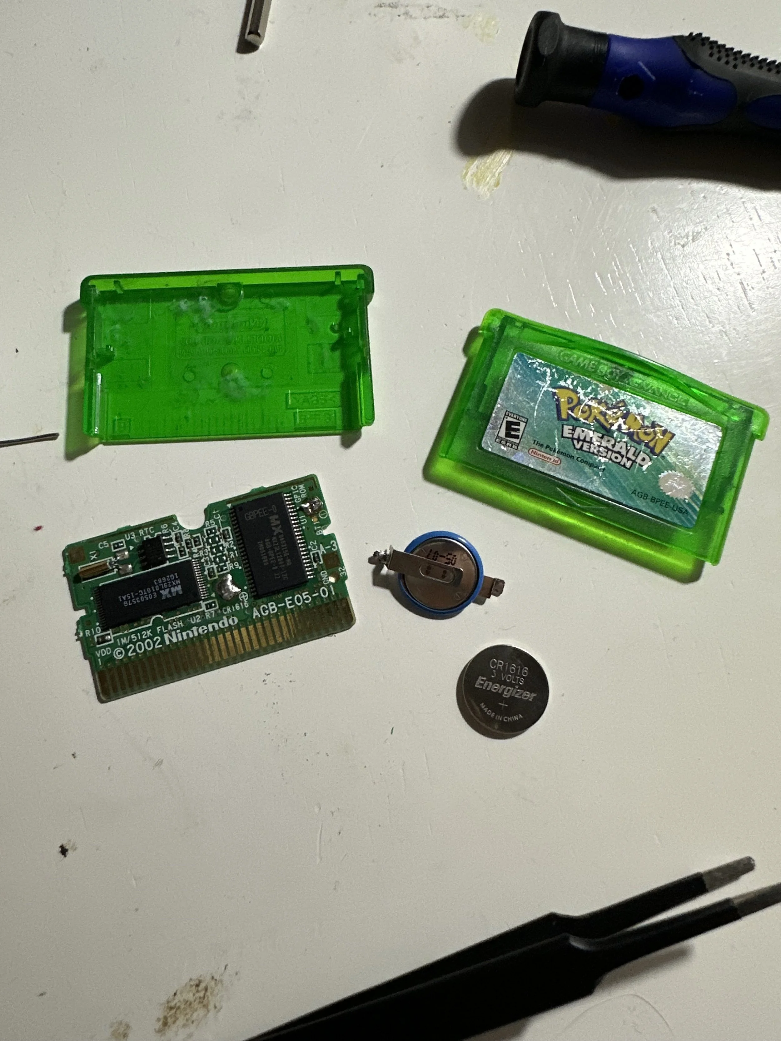 Disassembled electronic items including a green game cartridge labeled 'Pokémon Emerald Version,' a circuit board, batteries, a screwdriver, and other miscellaneous small components on a white surface.
