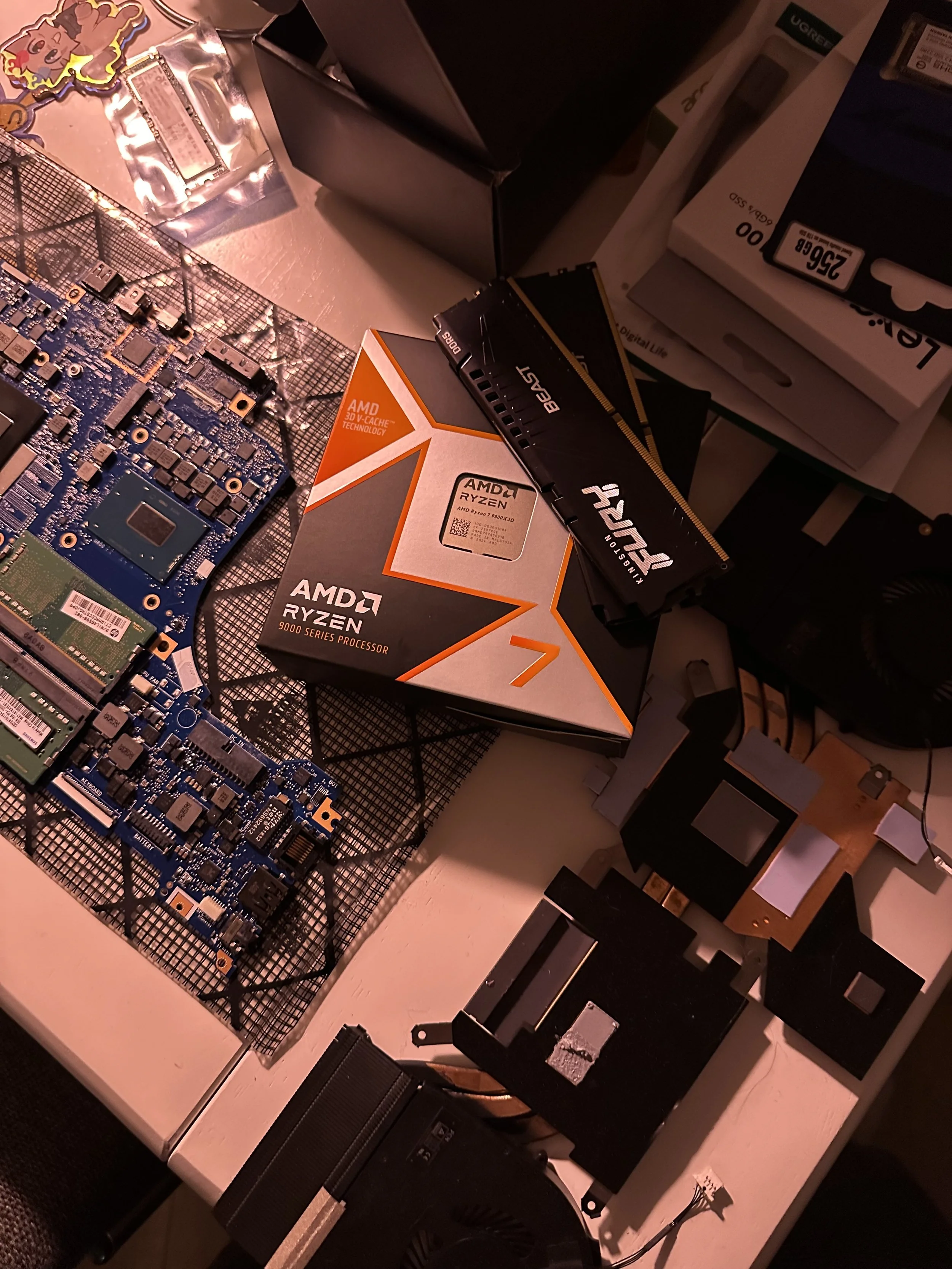A computer motherboard, a box of AMD Ryzen 9000 series processor, two RAM sticks, and various computer components and accessories on a desk.