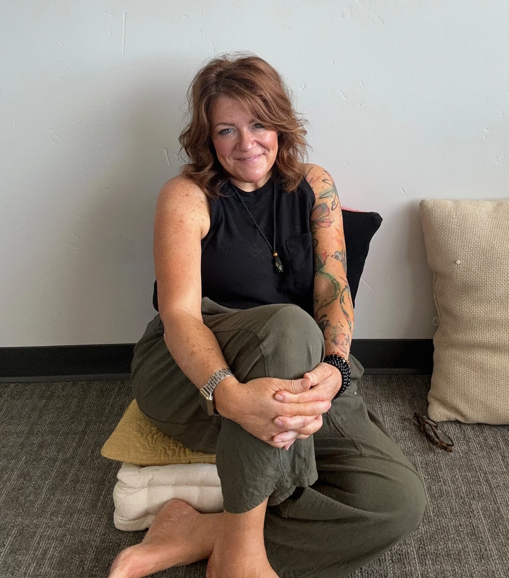 Woman with curly red hair, tattoos on her right arm, sitting cross-legged on a cushion with a smile. She is wearing a black top, jewelry, and bracelets, sitting indoors against a plain wall with a beige pillow nearby.