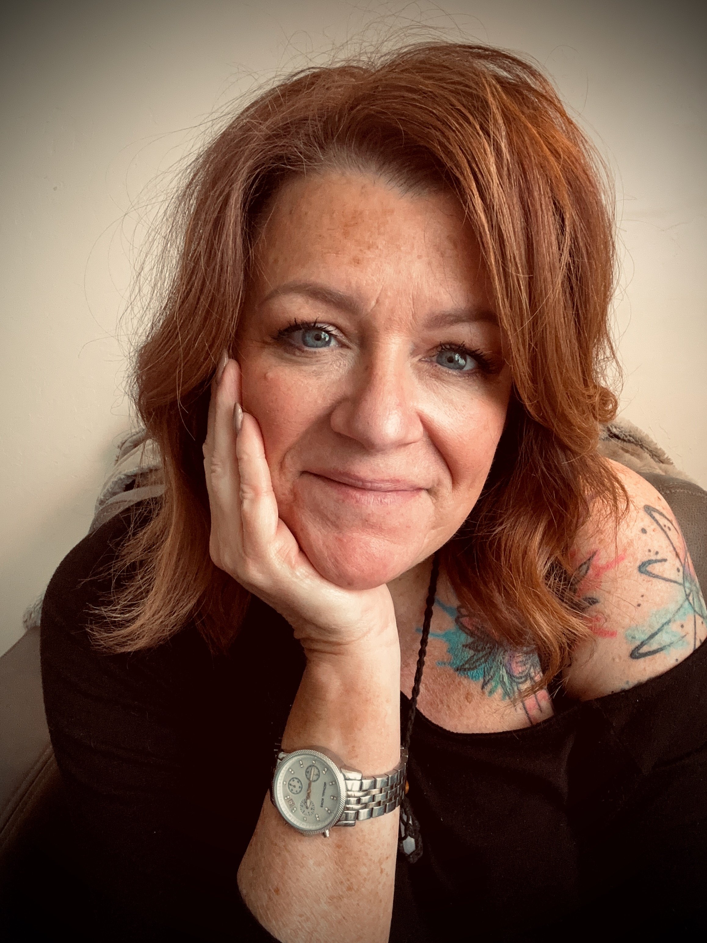 A woman with red hair, blue eyes, and tattoos on her arm, resting her head on her hand, wearing a silver watch and a black top, in front of a plain wall.