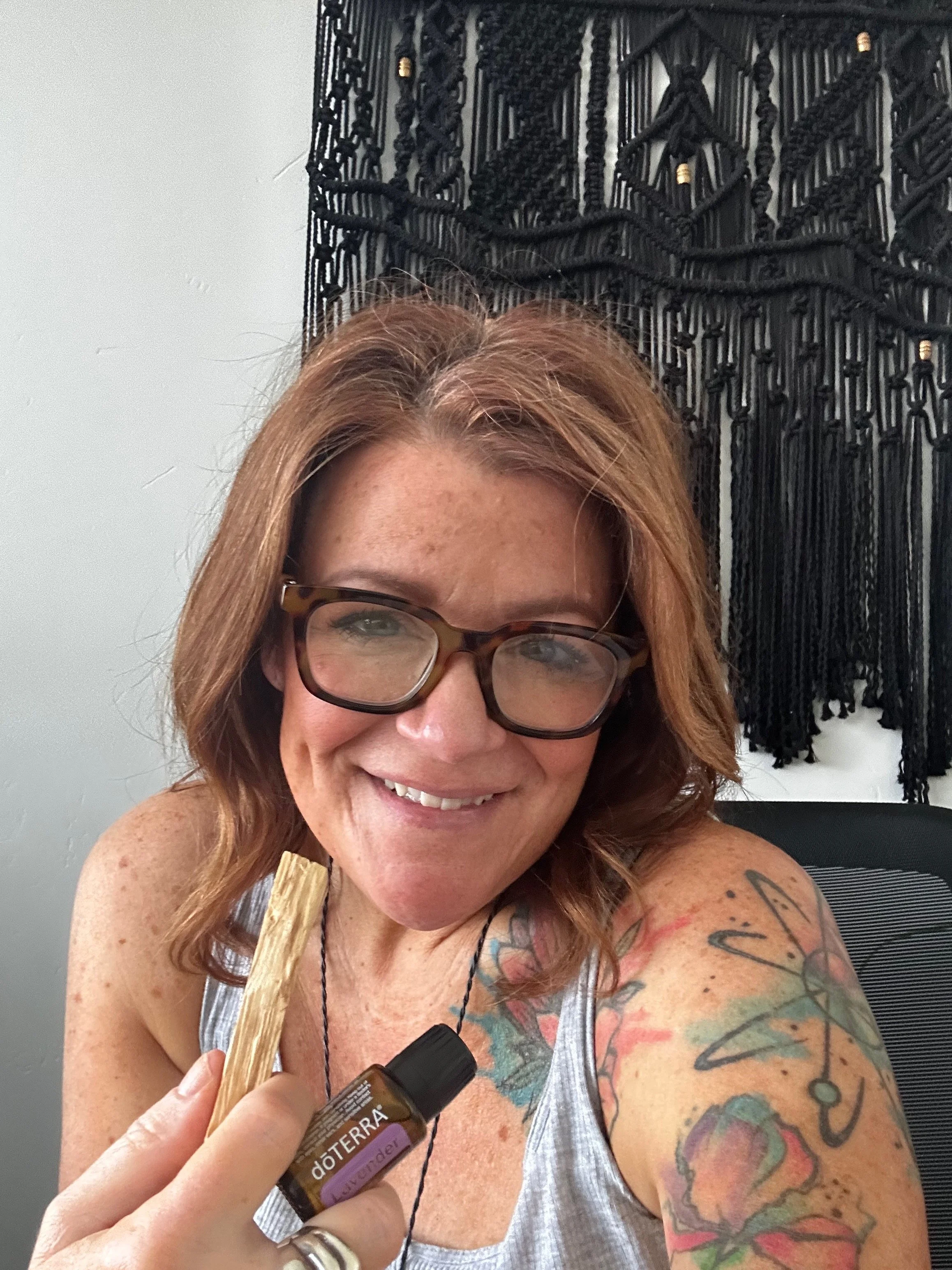 A woman with glasses and red hair smiling while holding a small bottle of doTERRA lavender essential oil. She shows her tattooed arm and a background piece of black wall art.