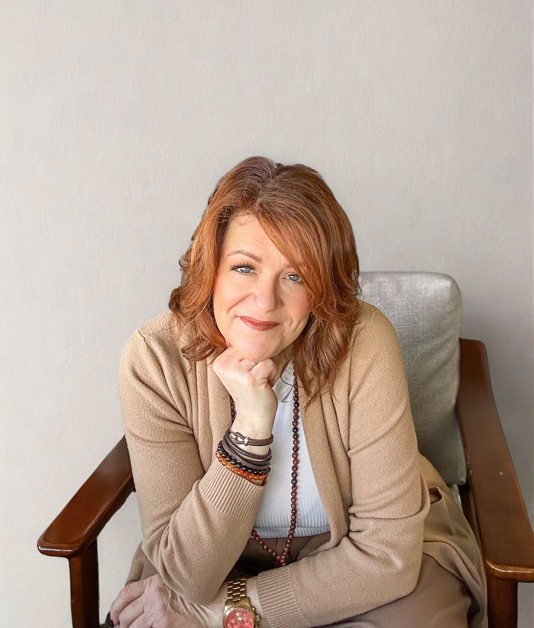 A woman with red hair and blue eyes sitting on a wooden chair with armrests, smiling with her hand resting on her chin, wearing a beige cardigan, white top, and multiple bracelets and a watch.