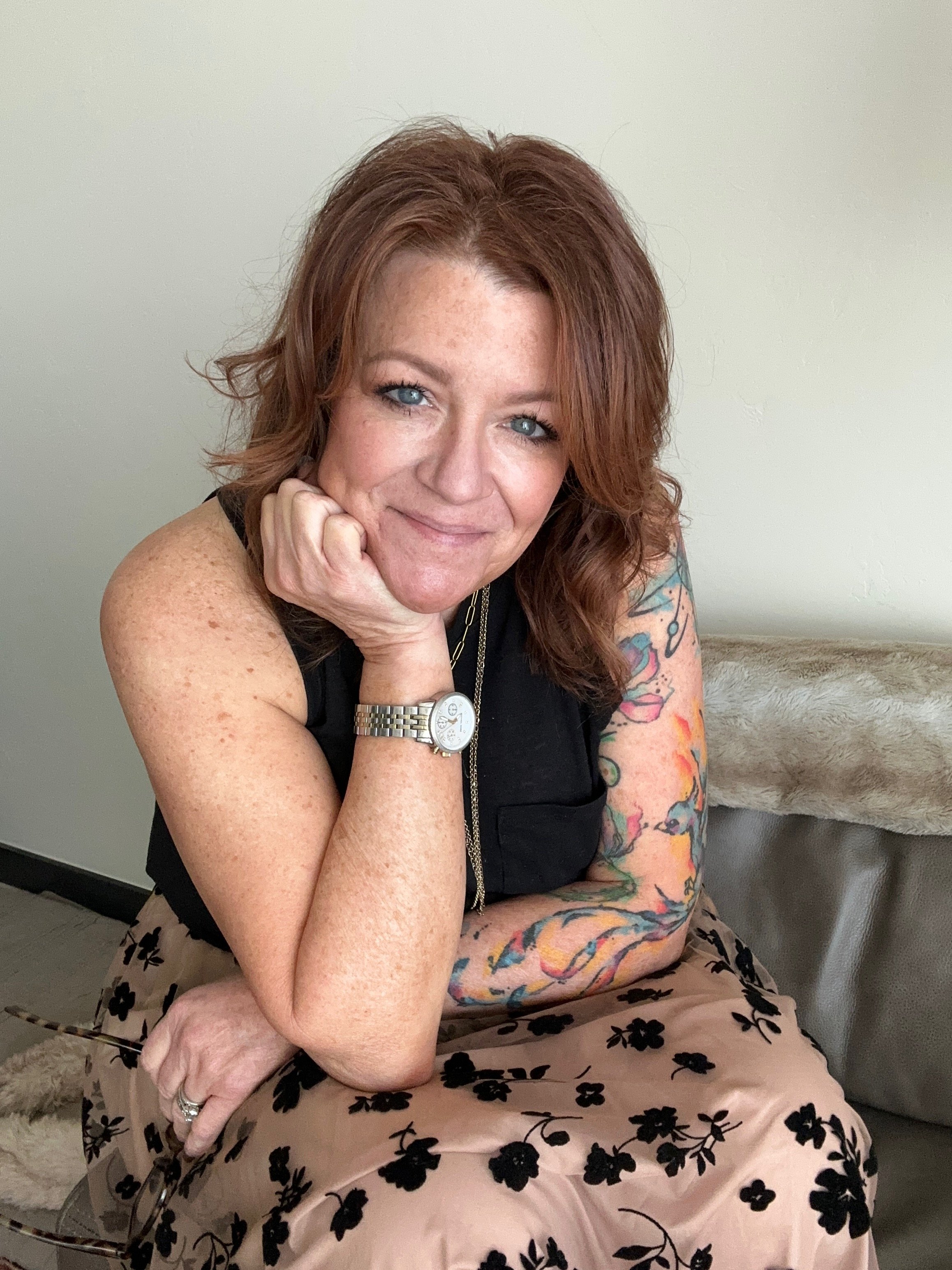 A woman with red hair and tattoos on her arms, wearing a watch and rings, seated with her elbow on her knee and hand supporting her chin, smiling at the camera against a plain light-colored wall.
