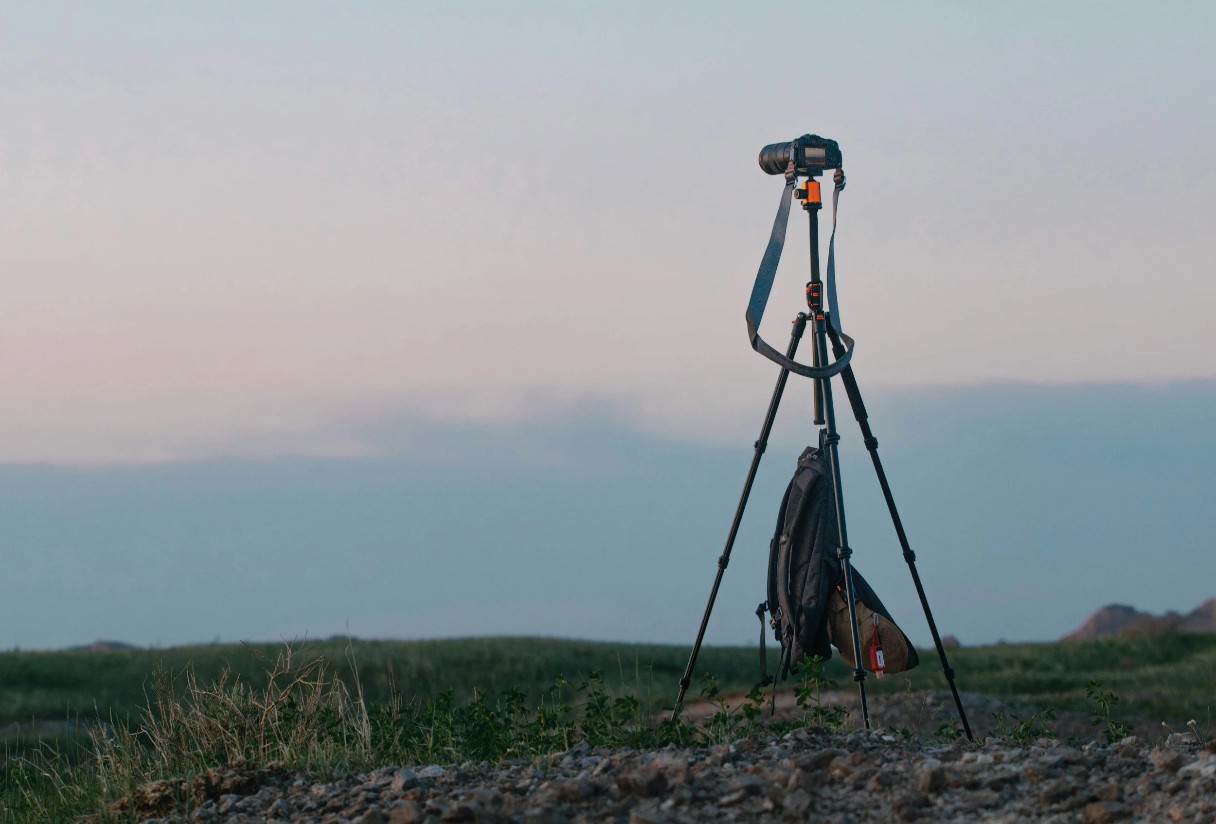 A camera mounted on a tripod stands in a field with a grassy landscape, against a backdrop of a pastel-colored sky with soft clouds at dusk or dawn.
