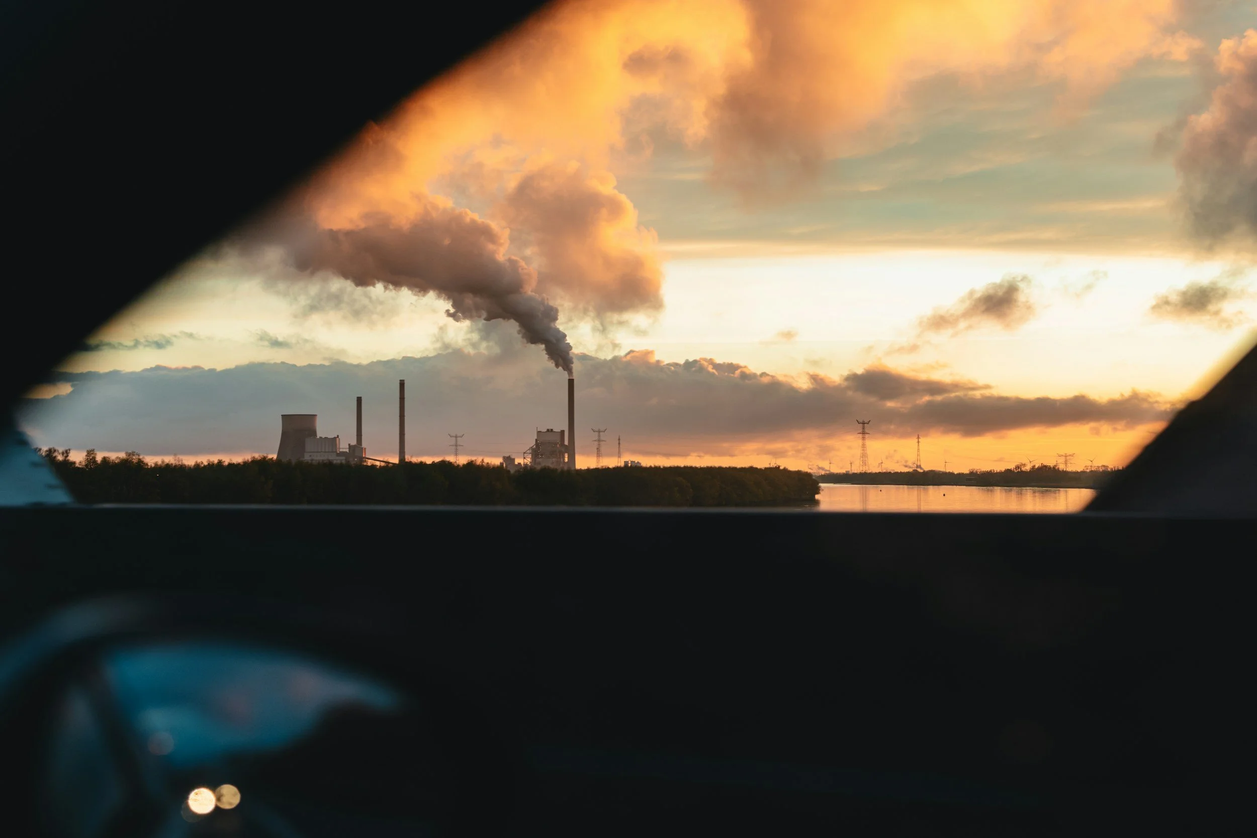 View of factory or power plant with smokestacks emitting dark smoke, seen through a vehicle window during sunset or sunrise with a partly cloudy sky.