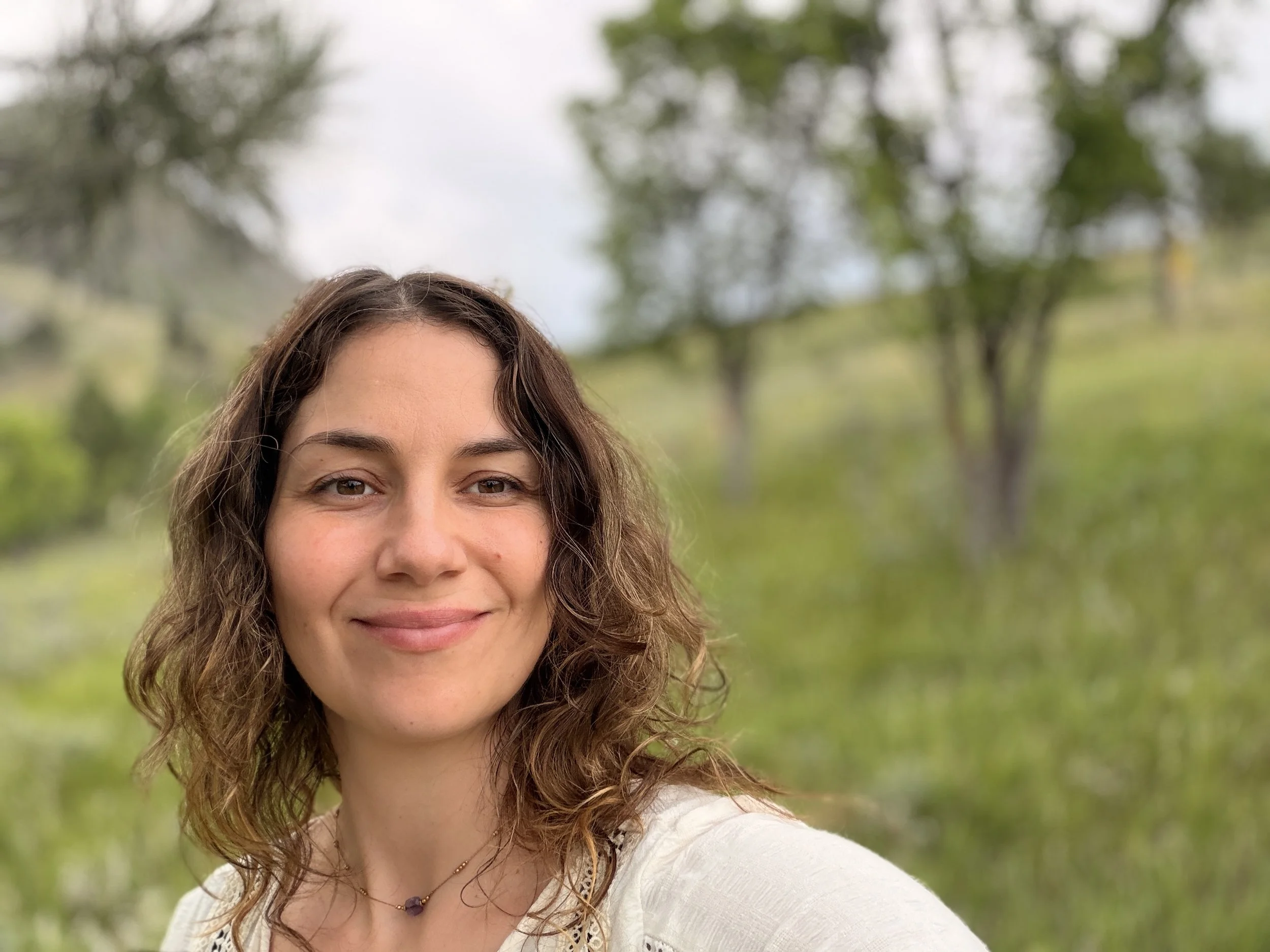 A woman with brown hair and brown eyes, smiling outdoors with greenery and trees in the background.