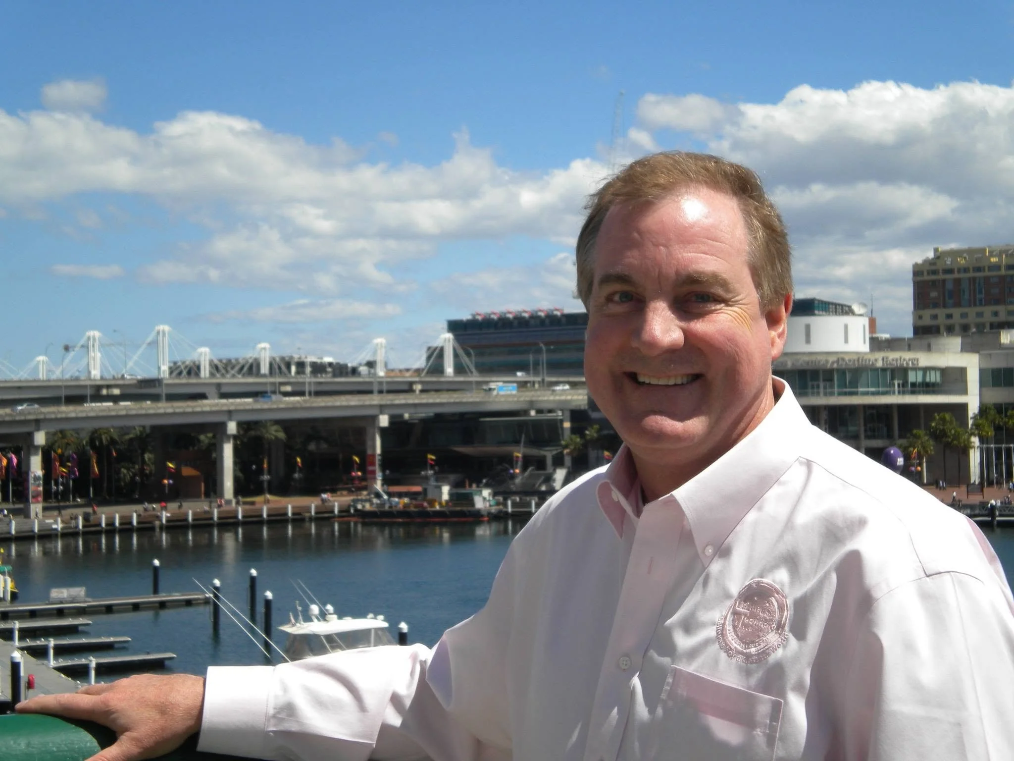 A smiling man in a white shirt standing near a marina with boats, a bridge, and buildings in the background under partly cloudy skies.