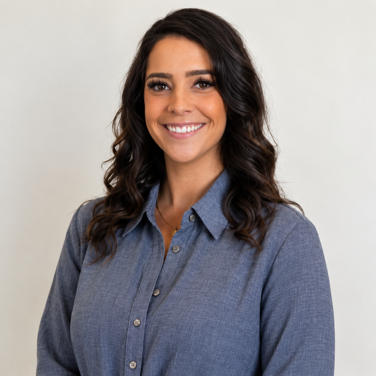 A woman with long wavy dark brown hair, smiling, wearing a gray button-up shirt, standing against a plain light-colored background.