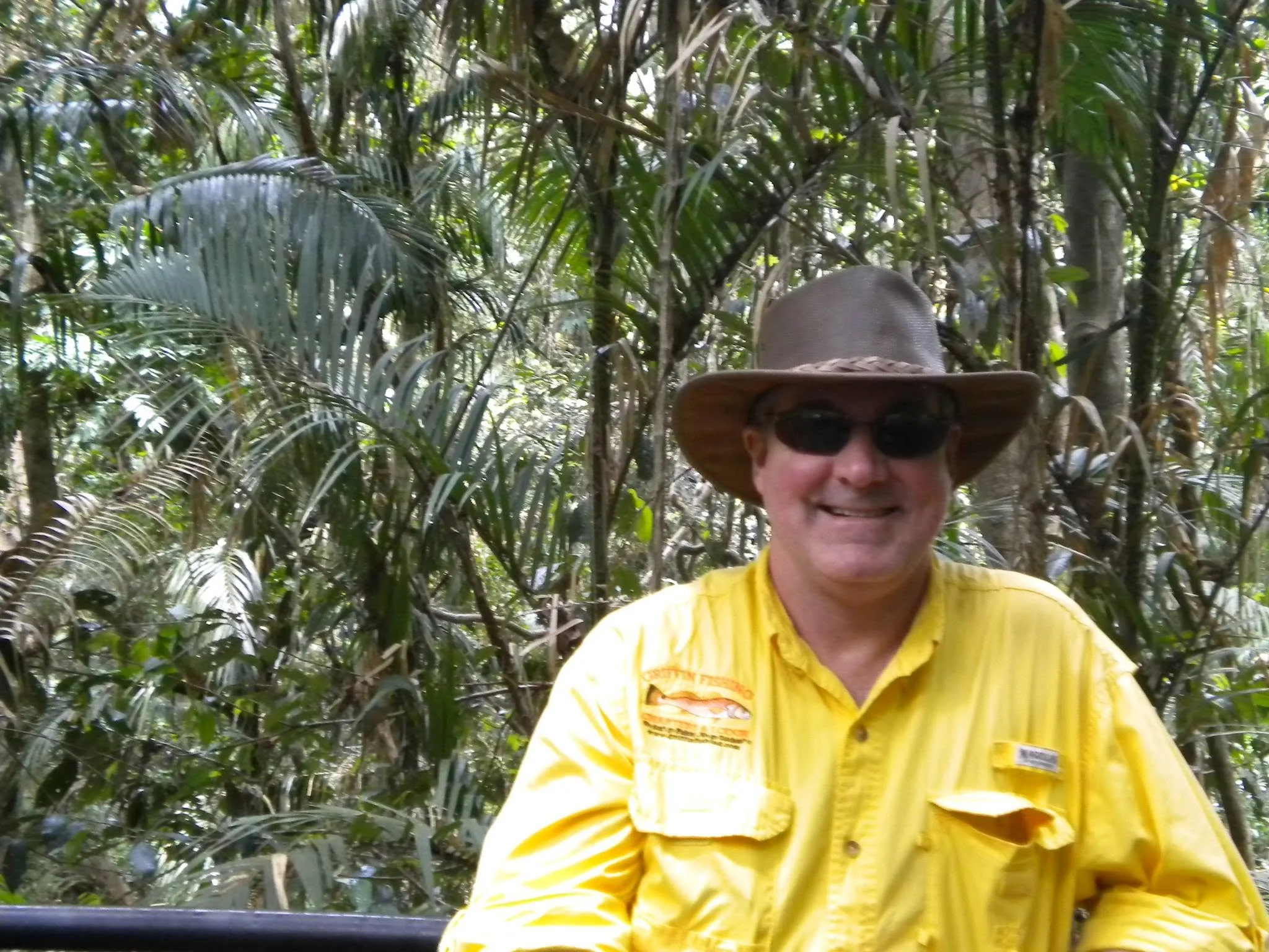 A person wearing a wide-brimmed hat, sunglasses, and a yellow shirt, standing in a dense jungle with lush green foliage surrounding him.