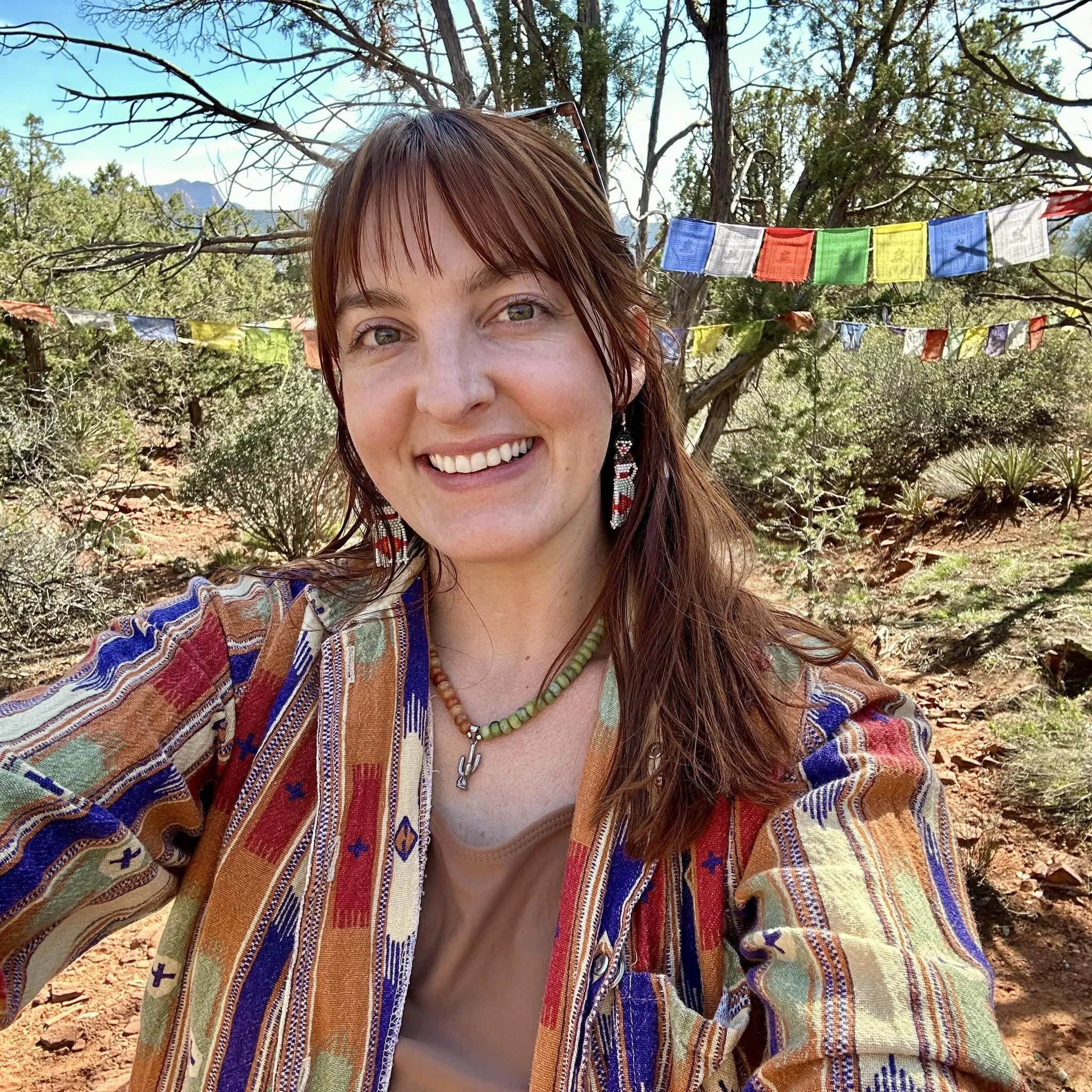 Close-up of a woman with reddish-brown hair smiling outdoors, wearing a colorful patterned jacket, a necklace, and earrings, with green trees, prayer flags, and mountains in the background.