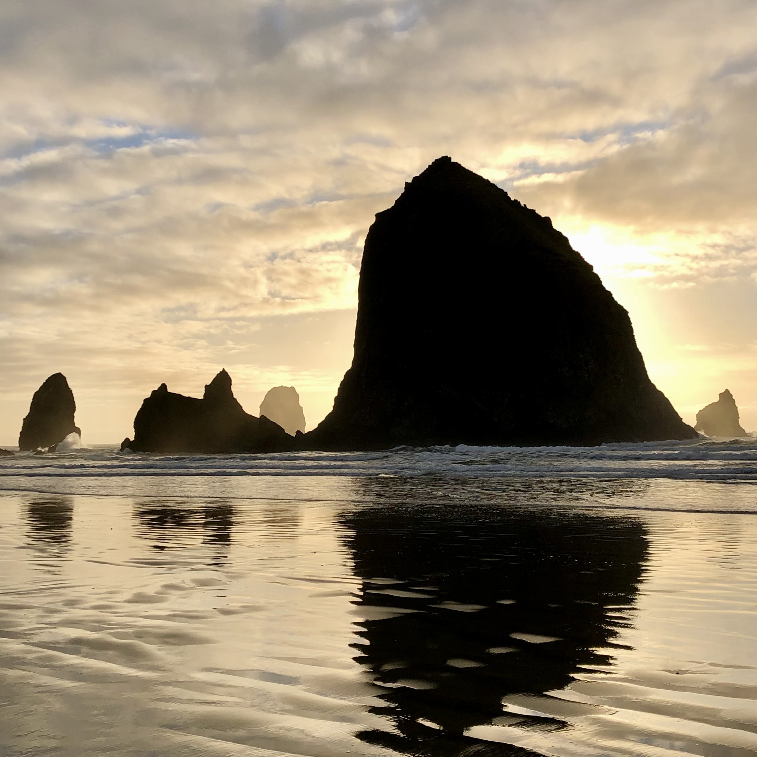Silhouetted rock formations on a beach at sunset or sunrise, with a partly cloudy sky reflected in the water.