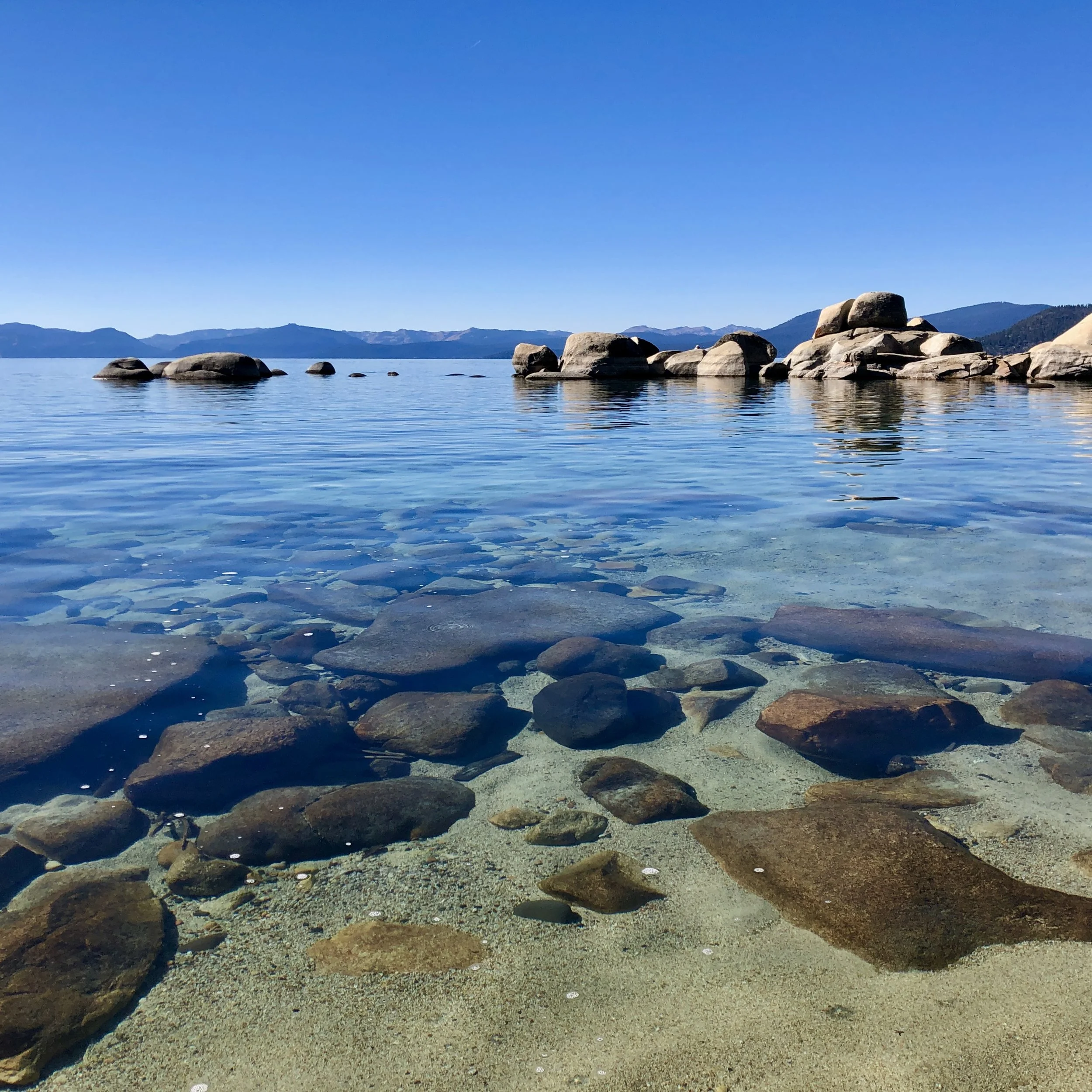 A calm lake with large rocks near the shore and mountain ranges in the background under a clear blue sky.