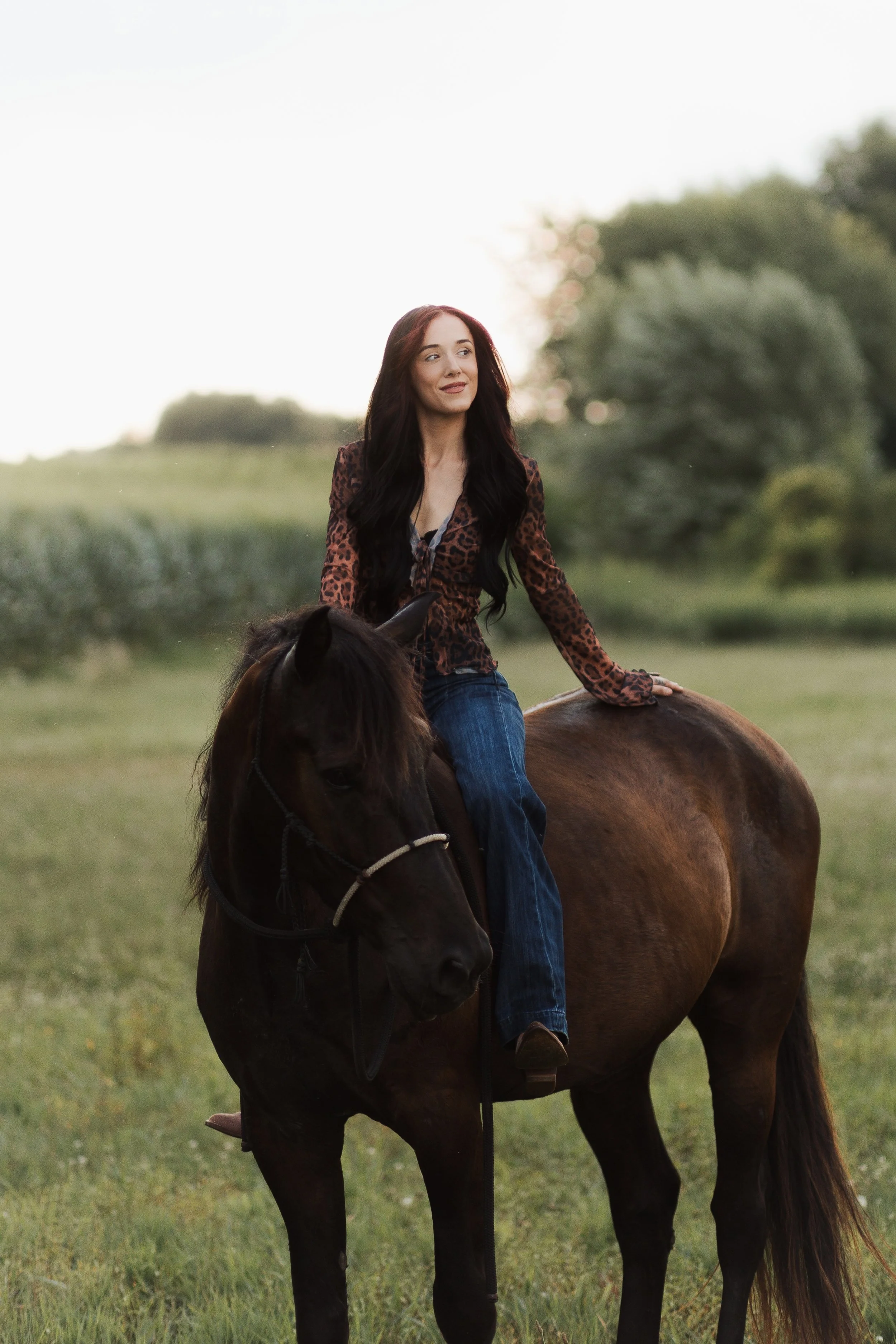 A woman with long hair riding a brown horse in a field during sunset.