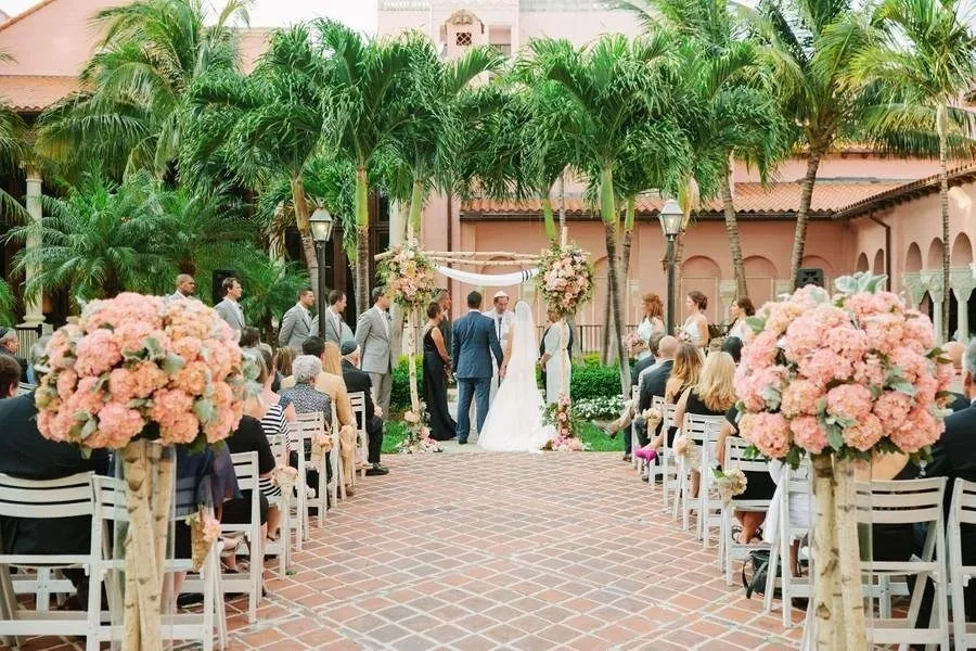 A wedding ceremony outdoors with palm trees, pink floral arrangements, and guests seated on white chairs.