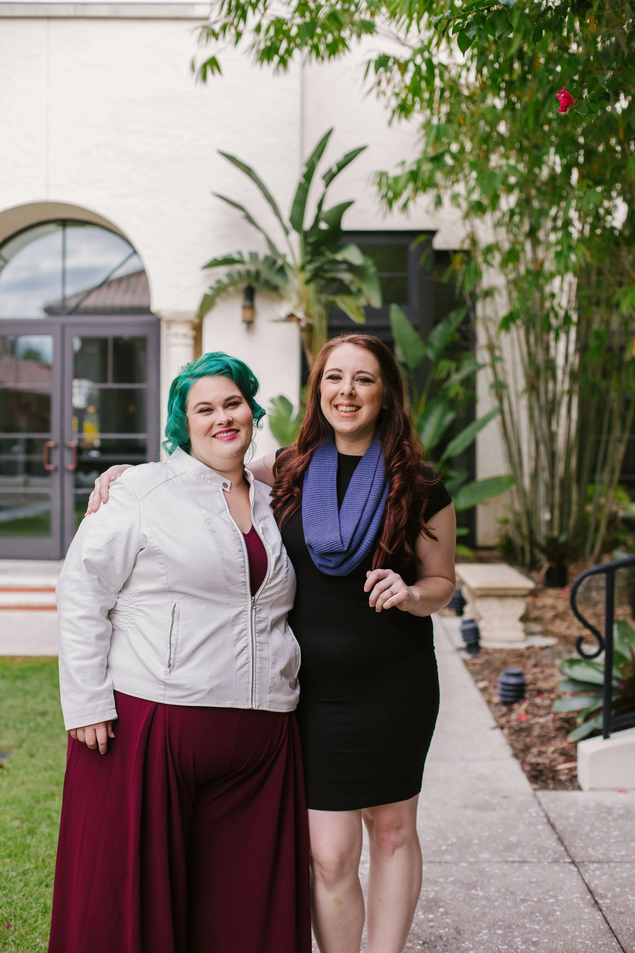 Two women standing outside in a courtyard, smiling and posing for a photo. On the left, a woman with green hair smiles in a burgundy dress and white jacket. On the right, a woman with reddish-brown hair wears a black dress and a blue scarf.