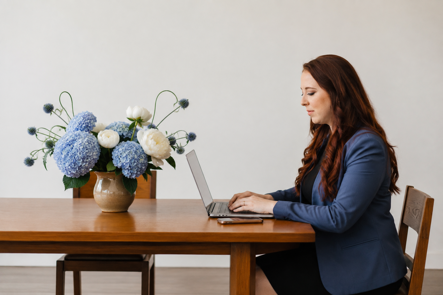 A woman with long, wavy red hair wearing a blue blazer, sitting at a wooden table, working on a silver laptop with a smartphone beside her. There is a large bouquet of blue and white hydrangeas in a beige ceramic vase on the table.