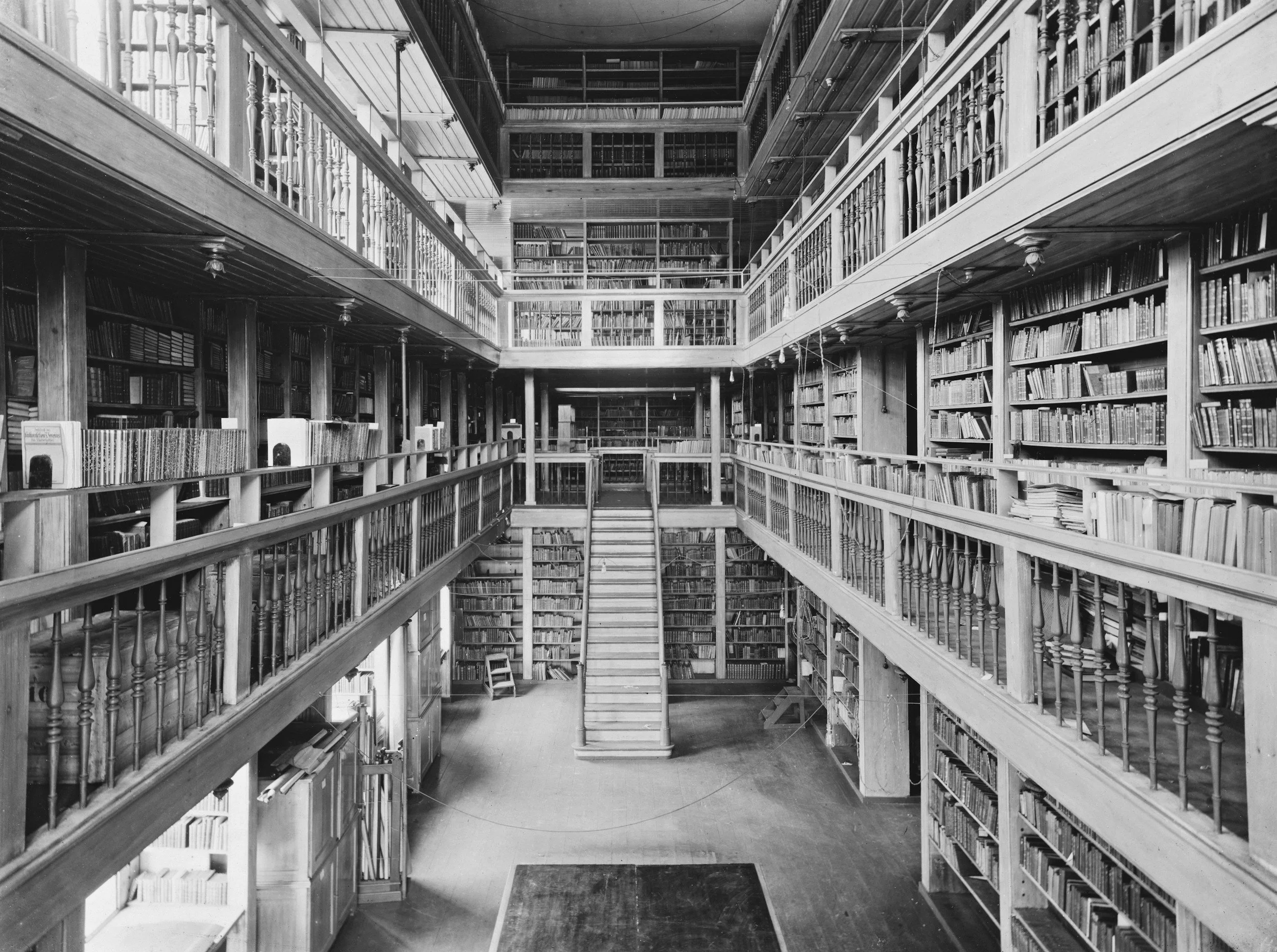 Black and white photo of a large multi-story wooden library interior with bookshelves lining the walls, balconies, and a staircase leading to upper levels.