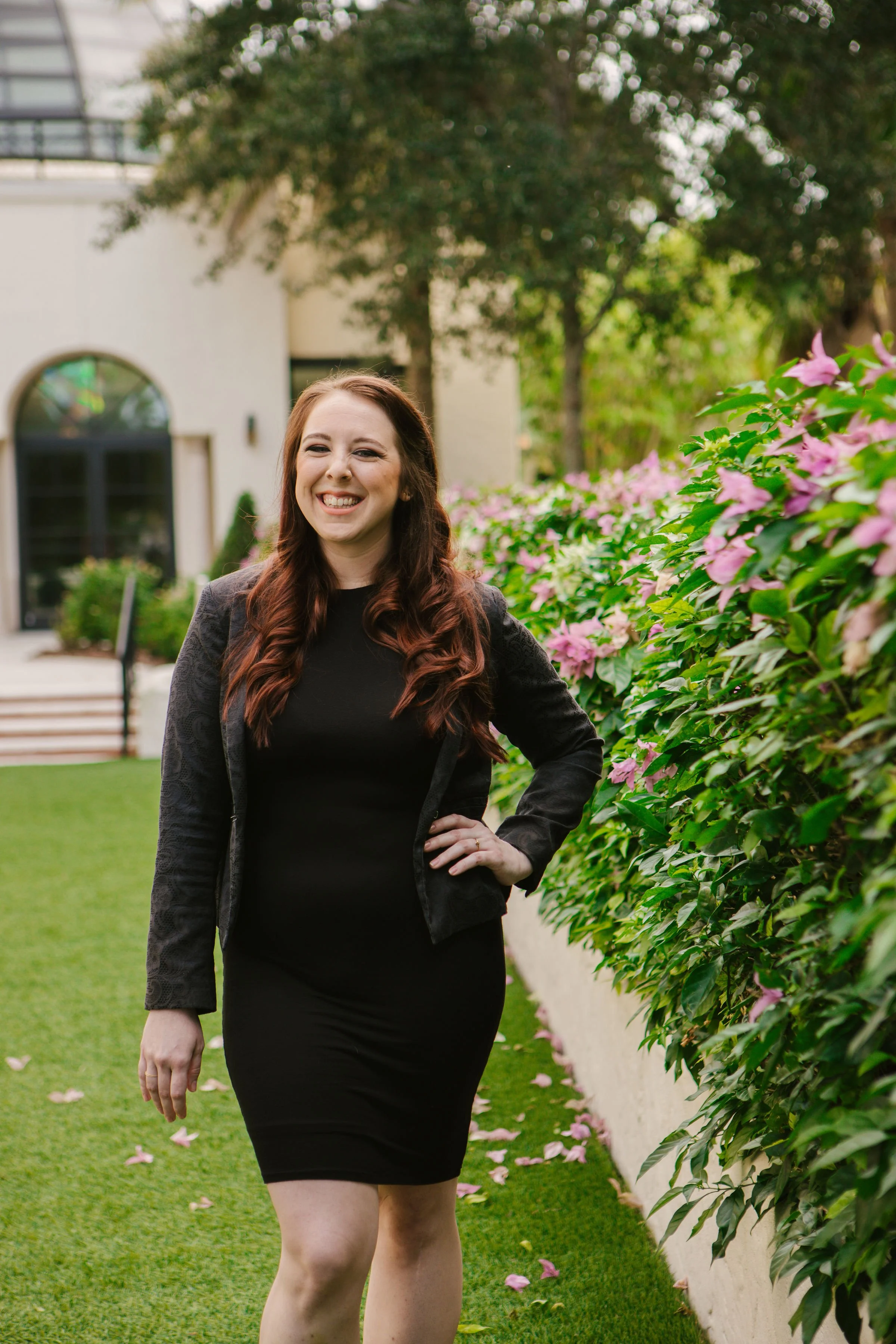 A smiling woman with long, wavy, reddish-brown hair wearing a black dress and a black lace blazer walking along a garden pathway with pink flowers on the right side and a modern building with large windows in the background.