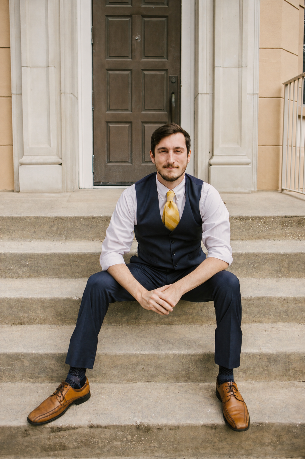 A professional man in a navy suit, white shirt, and yellow tie is sitting on stone steps in front of a brown door, with columns and railings of a nearby building visible.