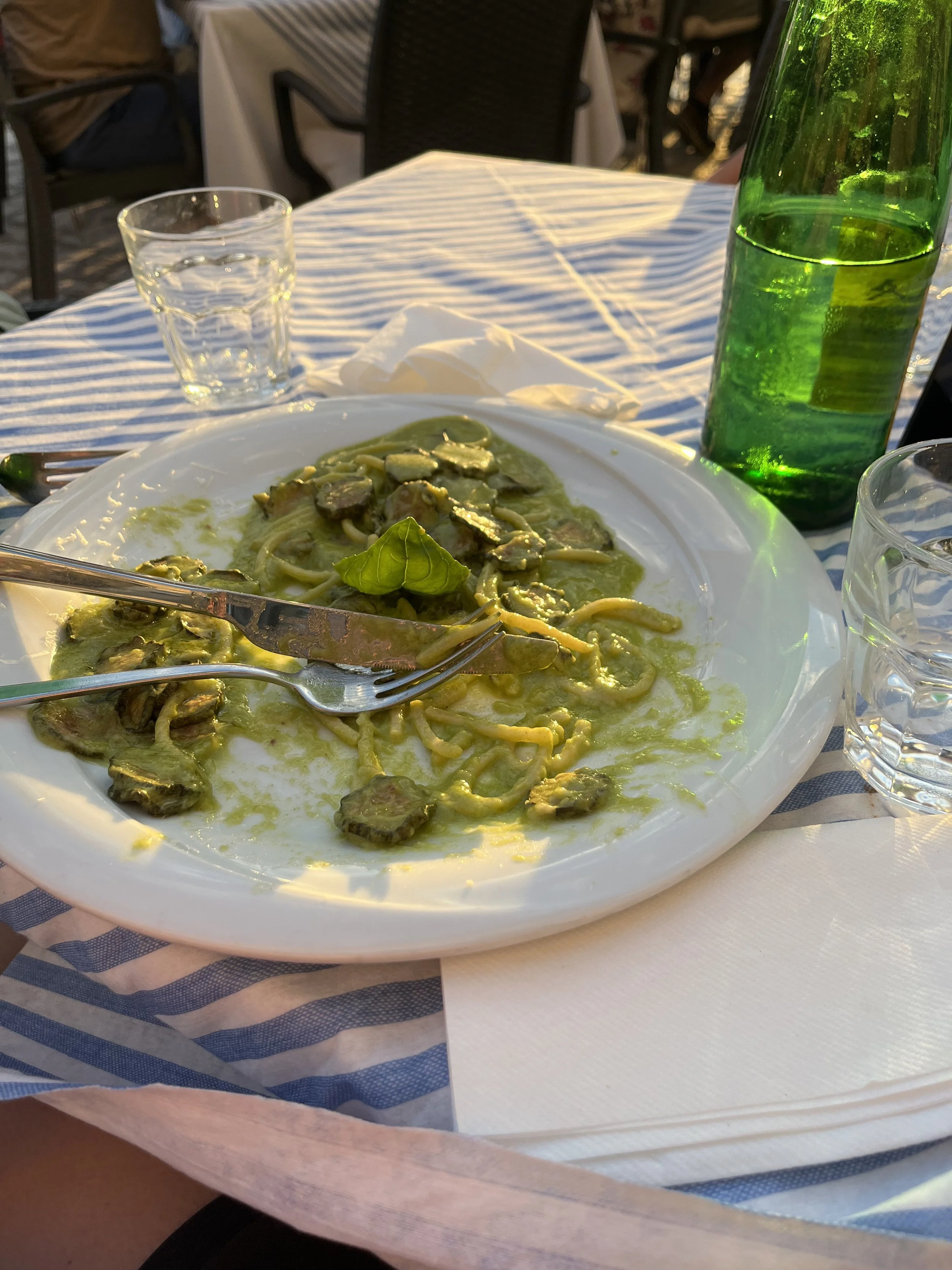 A plate of pasta with green sauce and sliced meat, on a table with a blue and white striped tablecloth, accompanied by a glass of water and a tall green bottle, outdoors at a restaurant.