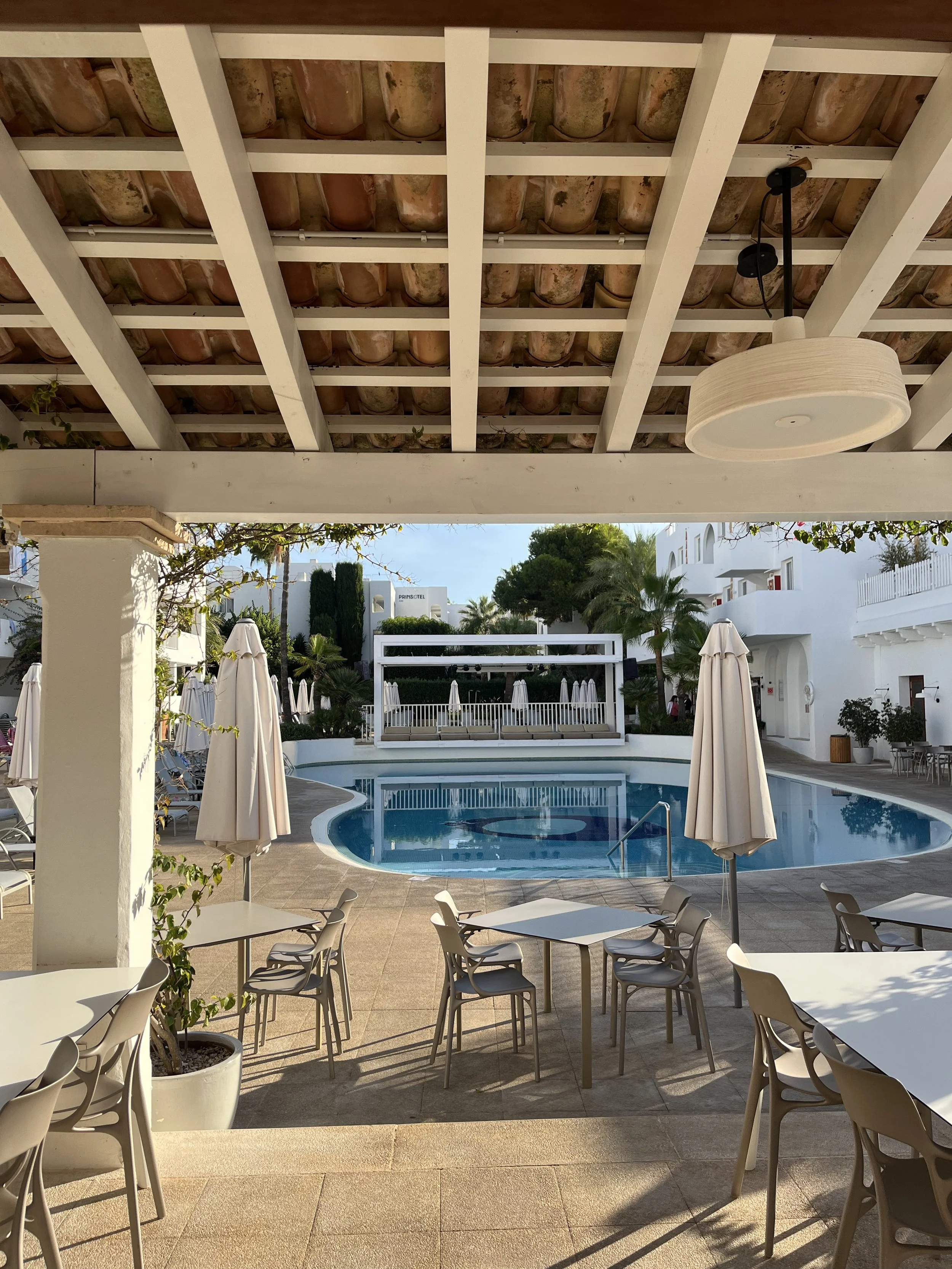 Outdoor hotel courtyard with a swimming pool, closed umbrellas, patio tables and chairs, palm trees, and white buildings.