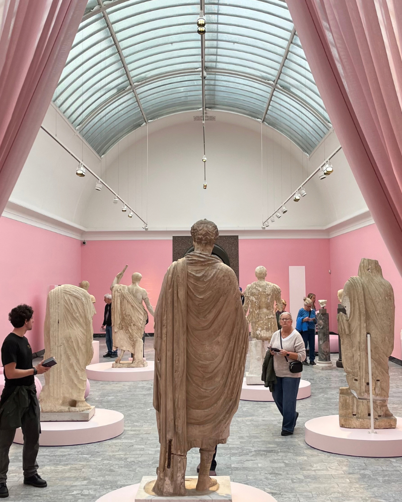 Visitors viewing ancient Greek and Roman marble statues in a pink-walled museum gallery beneath a glass-vaulted ceiling.