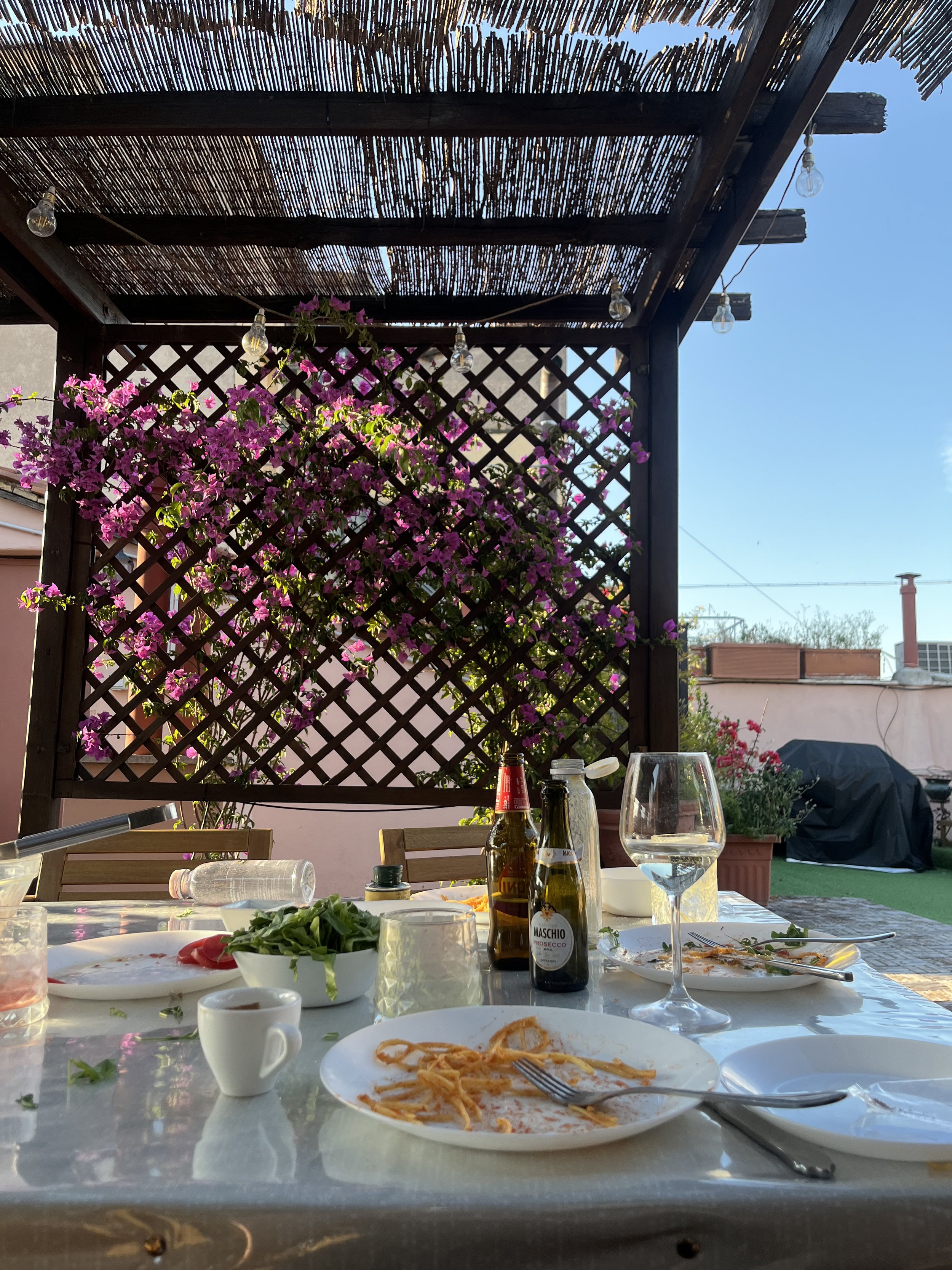 Outdoor dining table with leftover pasta, wine bottles, glasses, and plates beneath a reed pergola covered in blooming bougainvillea.