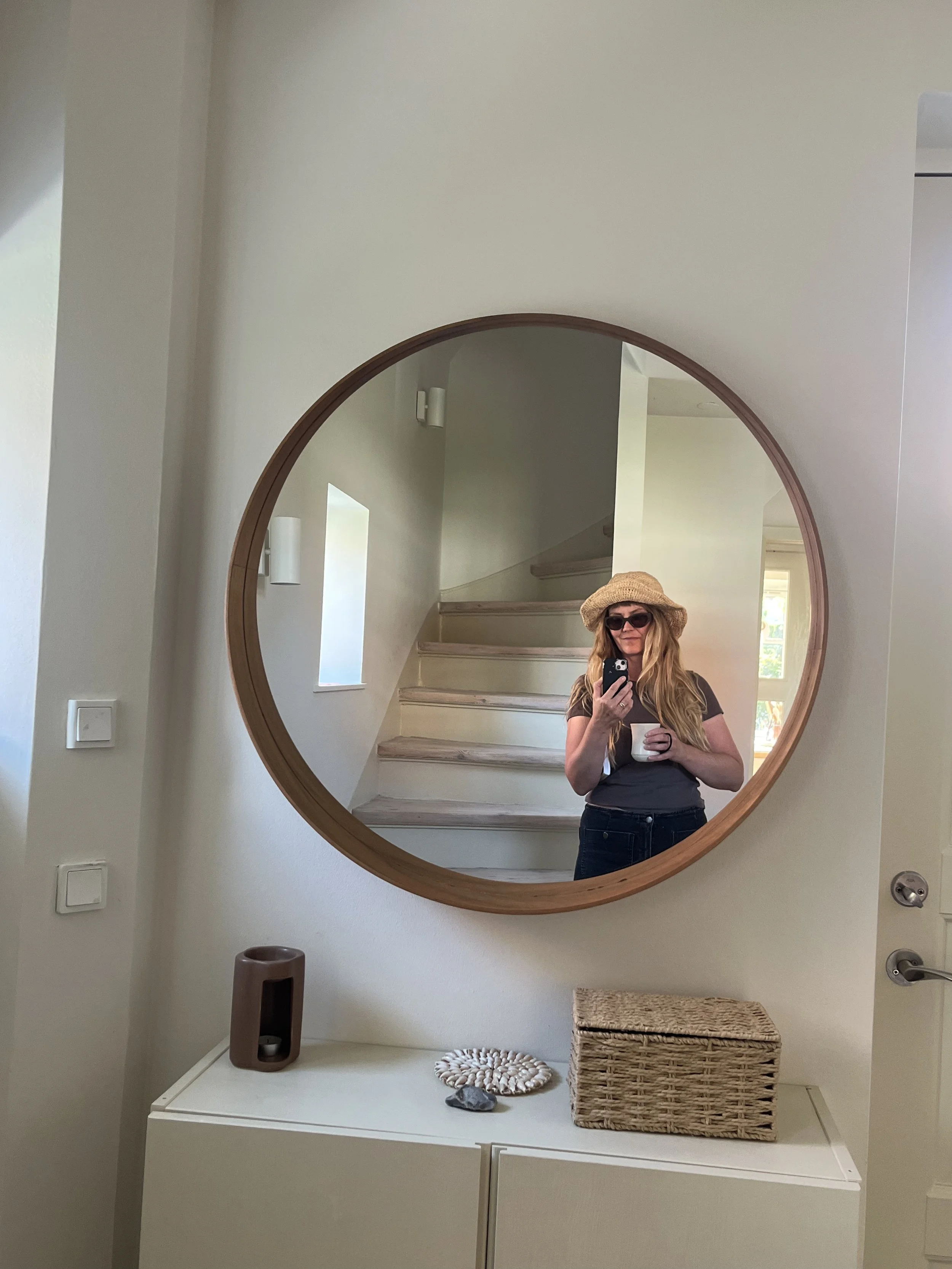 Woman wearing a straw hat and sunglasses takes a selfie in a round wood-framed hallway mirror while holding a cup.