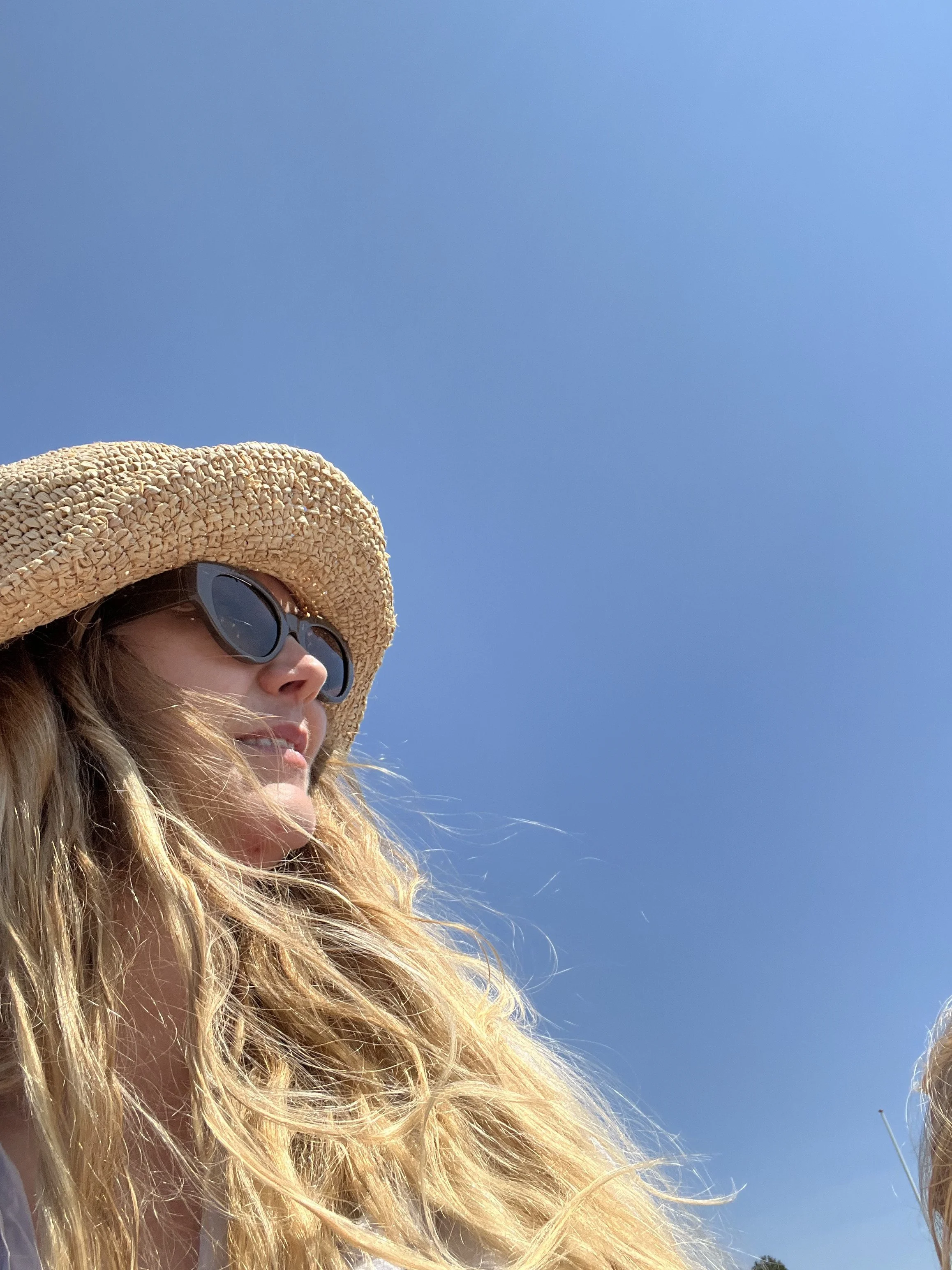 A woman with long, wavy blonde hair wearing a large straw hat and dark sunglasses under a clear blue sky.
