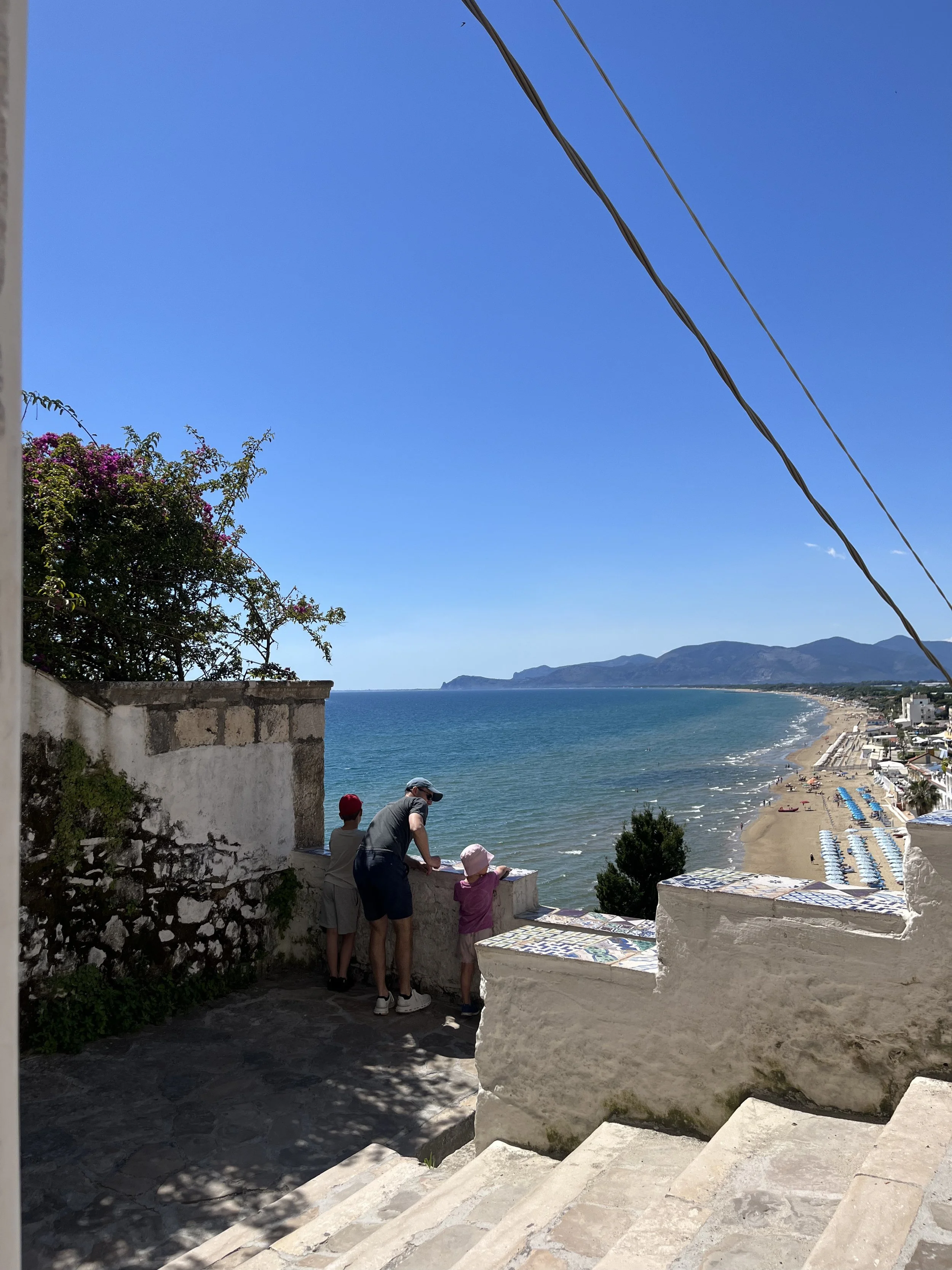 People standing beside a white stone wall overlooking a beach and ocean, with mountains in the distance under a clear blue sky.