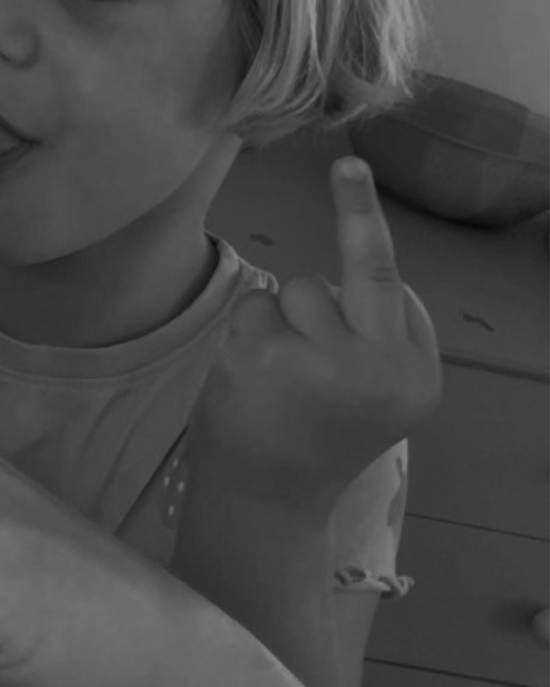 A young person with light-colored hair making a rude gesture by raising their middle finger. They are wearing a wristband and a t-shirt, sitting indoors.