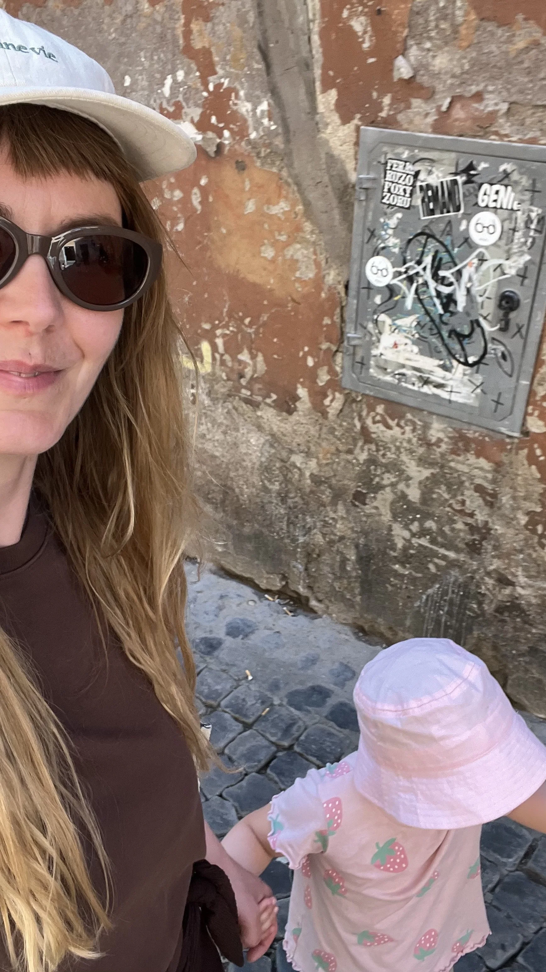 A woman wearing sunglasses and a white cap, standing next to a young girl in a pink dress with a strawberry print and a pink hat, holding hands on a cobblestone street next to a weathered brick wall with a graffiti-covered electrical box.