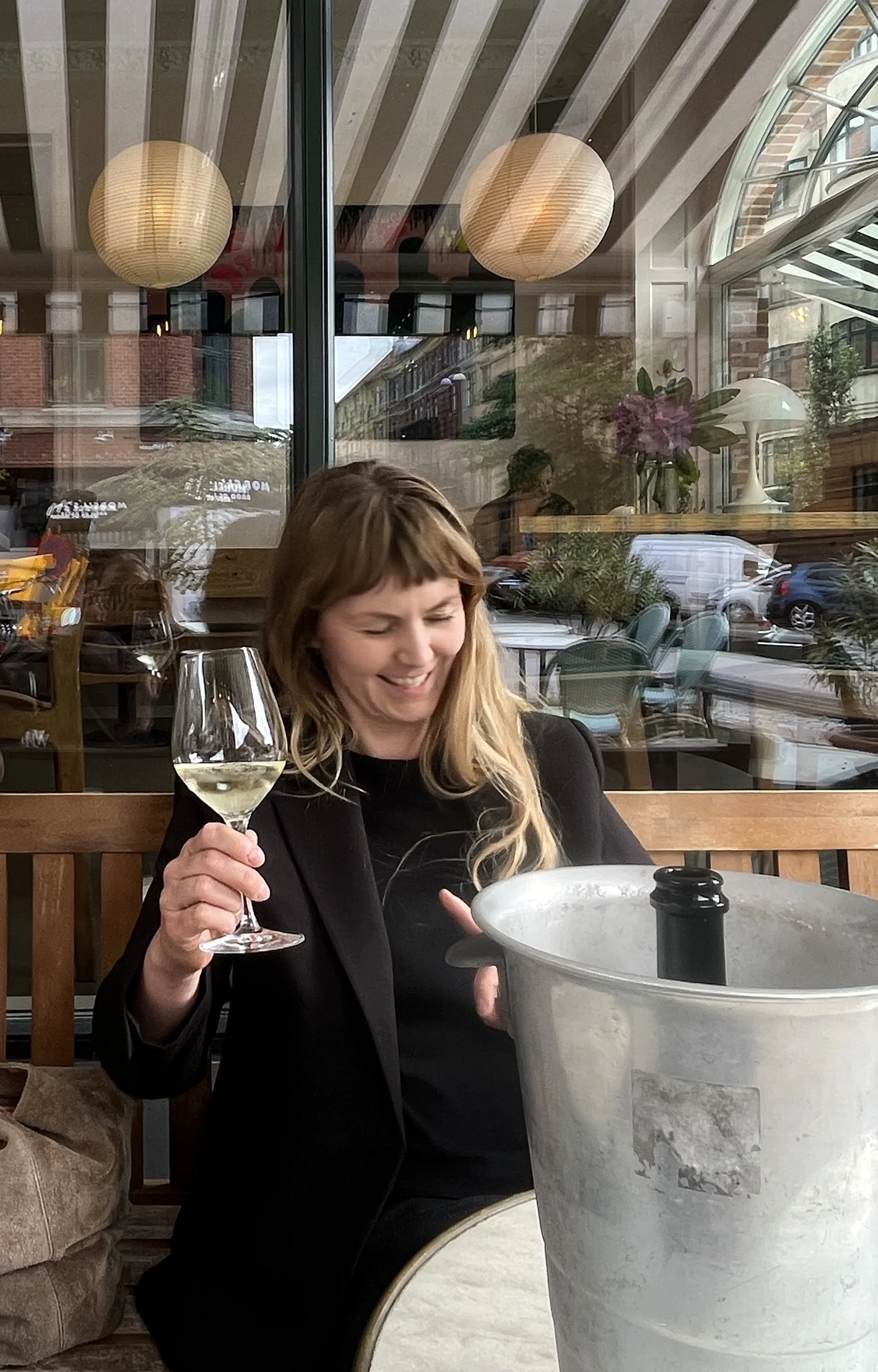 A woman with long blonde hair, wearing a black blazer, is sitting at a table in a restaurant or cafe, smiling and holding a glass of white wine. There is a large ice bucket on the table, and the background shows reflections of the street and interior lights through glass windows.