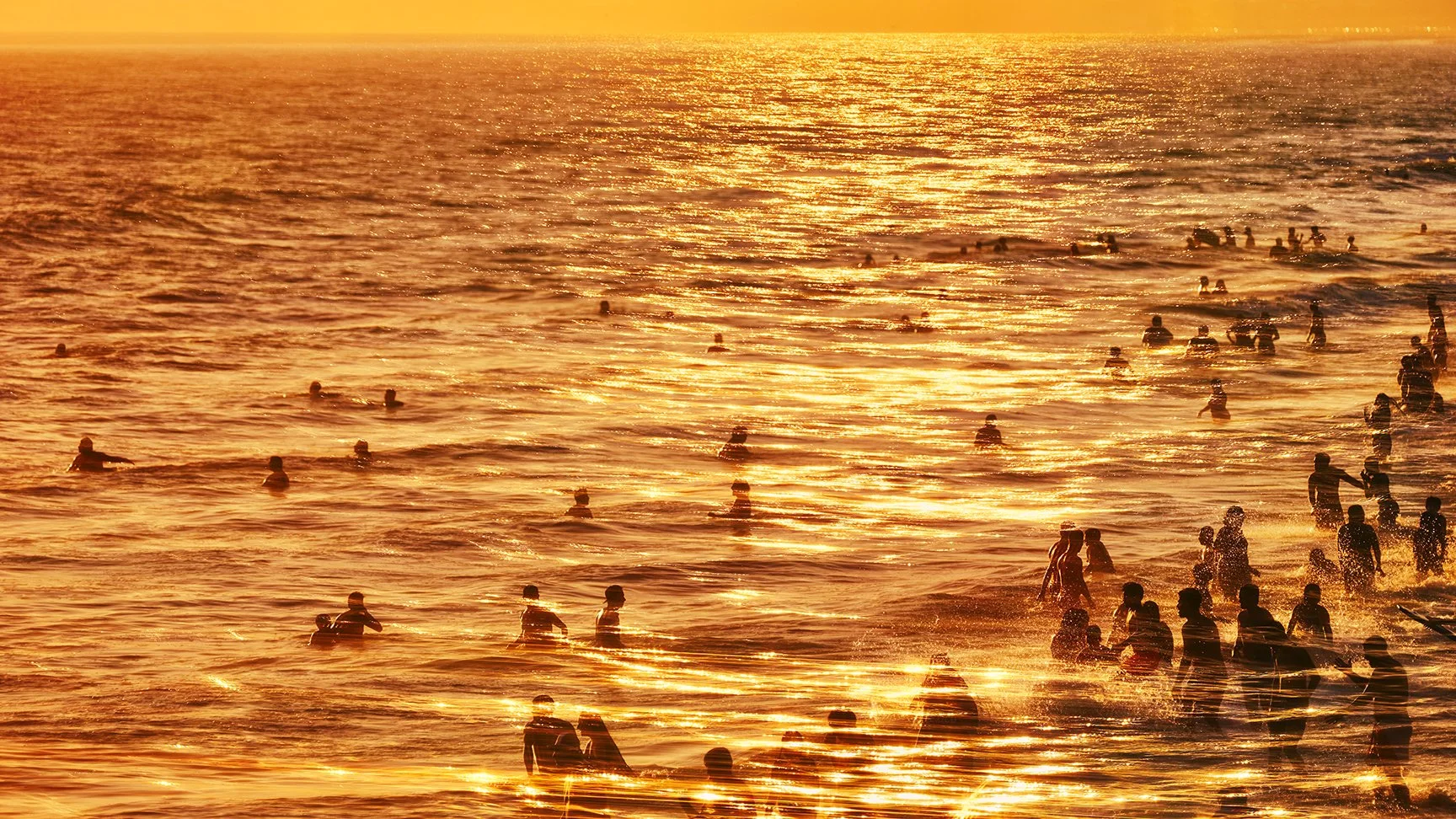 A sunset scene at the beach with many people swimming and playing in the water, illuminated by golden-orange sunlight.