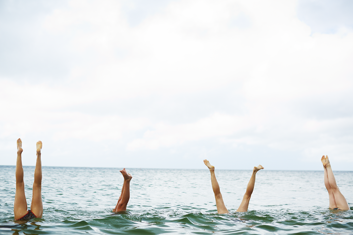 Four people are floating upside down in the ocean with only their legs and feet visible above the water, near a cloudy sky.