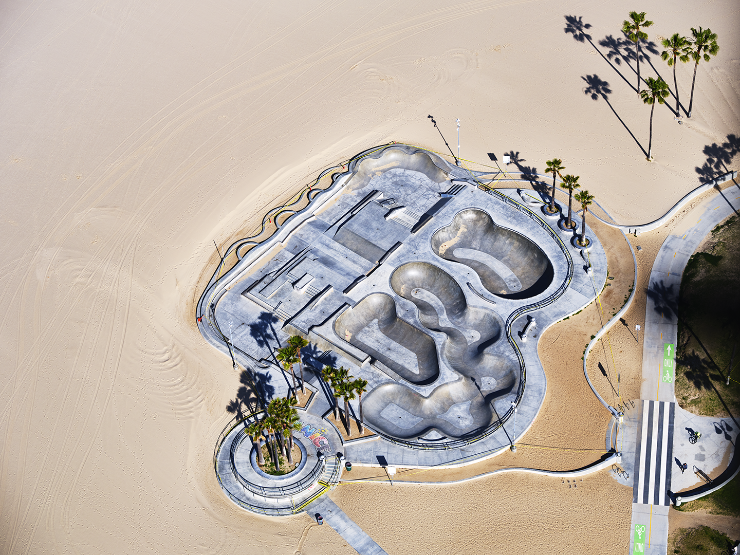 Aerial view of a concrete skate park with multiple bowls and ramps, surrounded by sand and a few palm trees, with a nearby bike lane and sidewalk.
