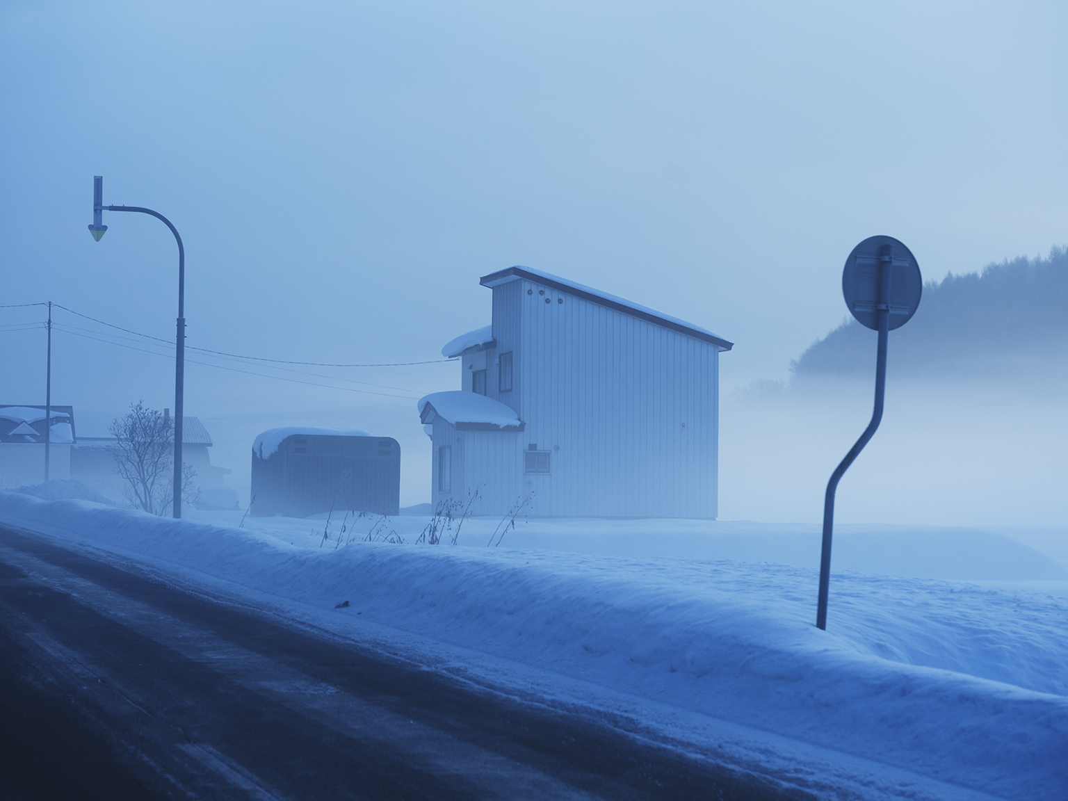 A snow-covered rural landscape with a small house, snow-laden trees, and street signs shrouded in fog or mist.