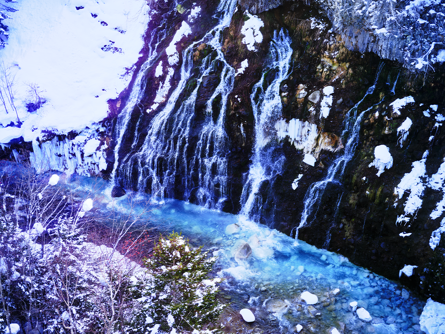 Frozen waterfall cascading into a river in a snowy, wooded landscape.