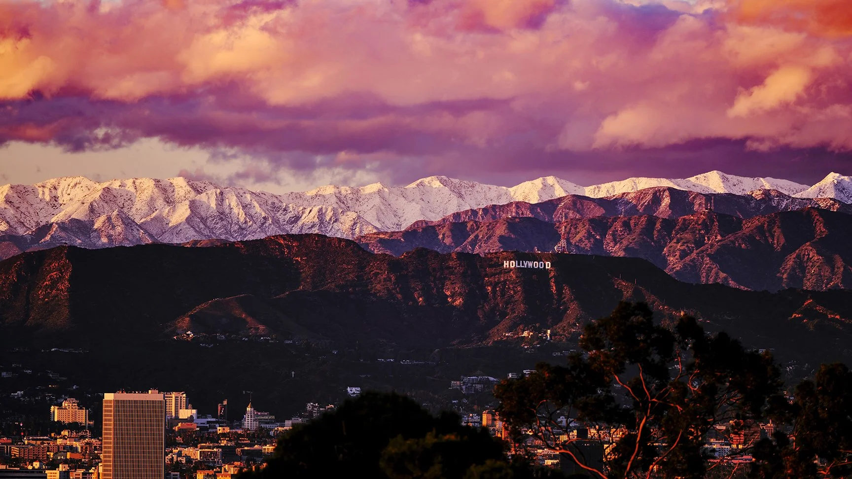Sunset view of the Hollywood sign on the hillside with city buildings in the foreground and snow-capped mountains in the background under a pink and purple sky.