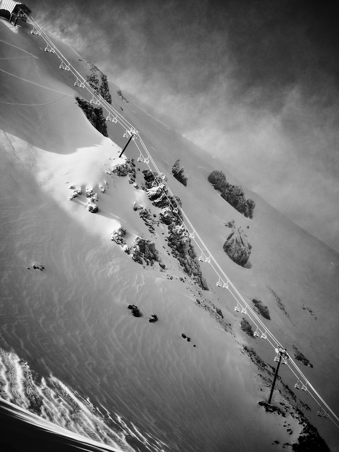Black-and-white photograph of a snowy mountain landscape with ski lift cables and chairs running diagonally across the image, rocky outcroppings, and trees on the slope.