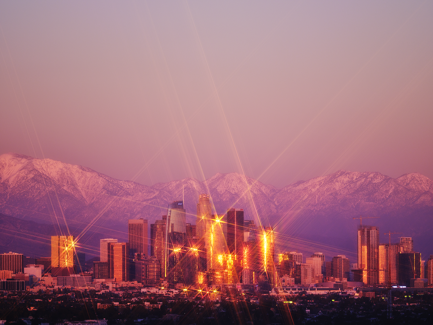 City skyline at sunset with tall buildings illuminated by streetlights, mountain range in the background, pink and purple sky above.