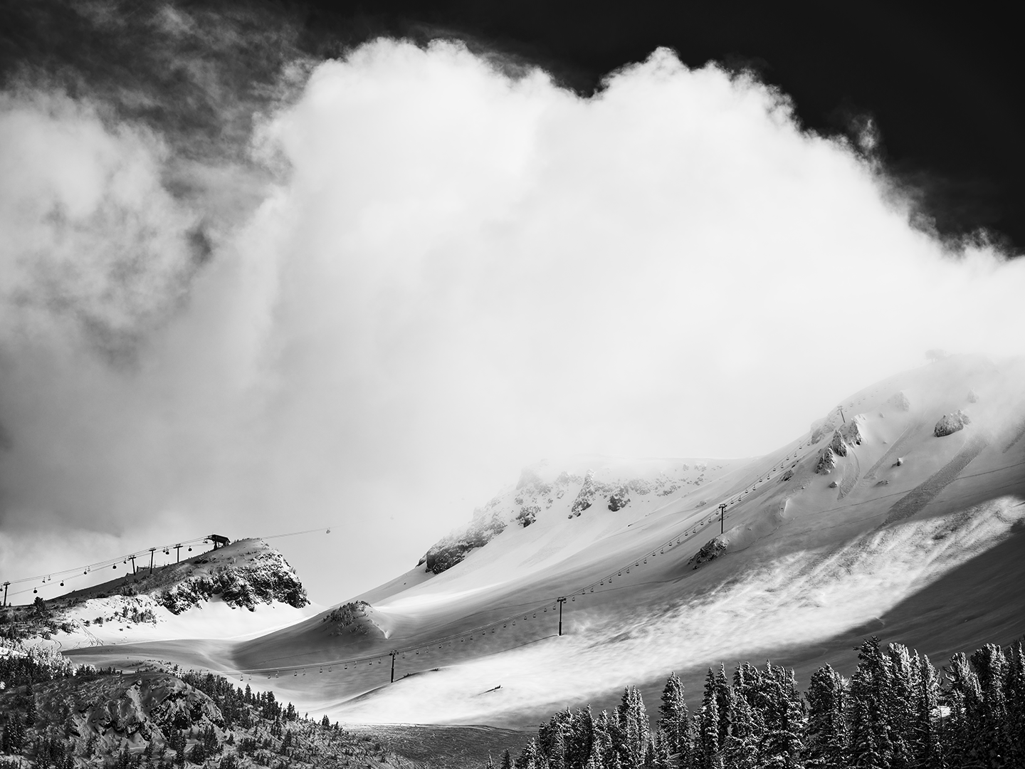 Snow-covered mountain landscape with ski lift lines and pine trees in the foreground, clouds above.