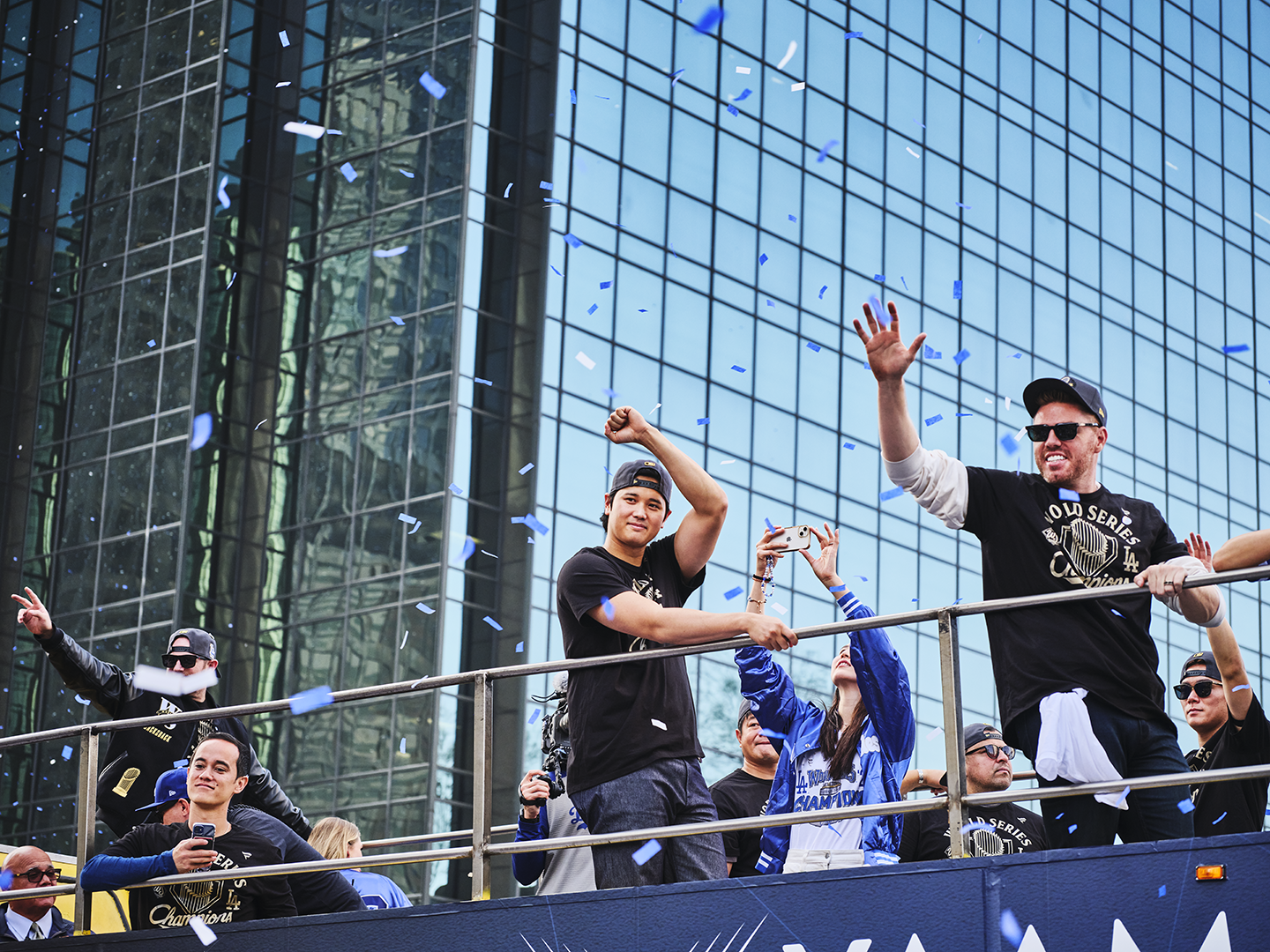 People celebrating on a parade float, some waving and taking photos, with blue confetti falling, in front of a modern glass building.
