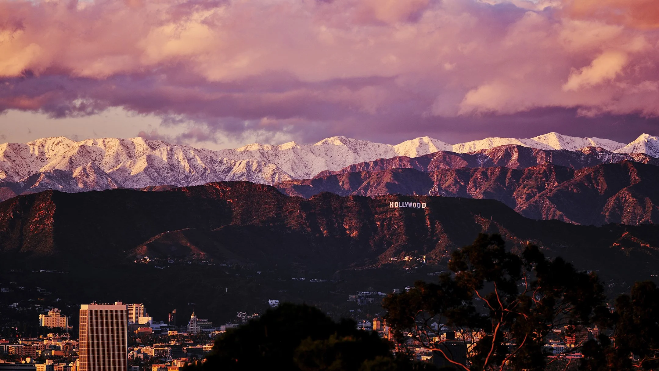 City skyline with tall buildings in front of snow-capped mountains at sunset, with a Hollywood sign visible on the mountainside.