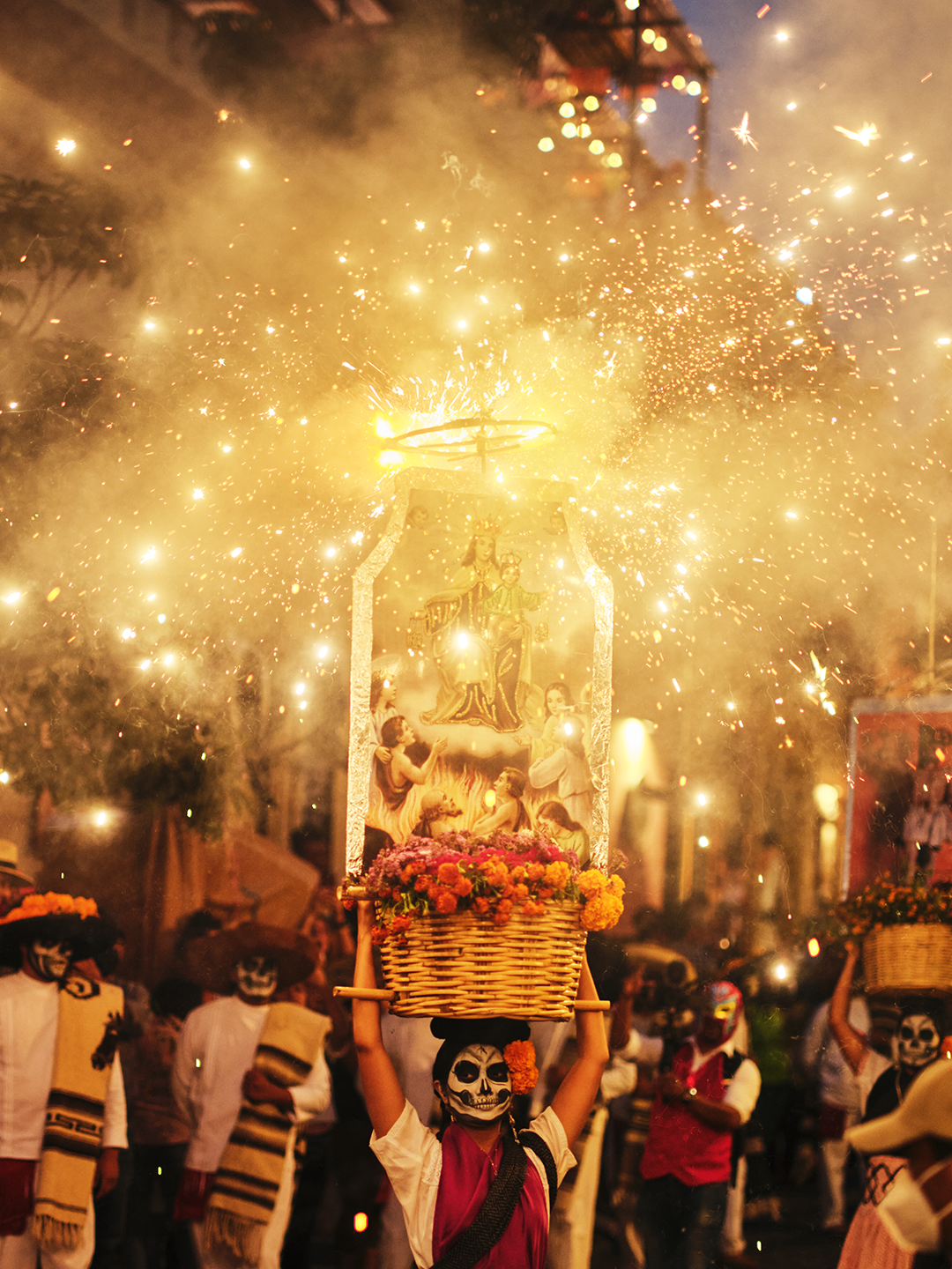 Person with painted face holding a basket with a religious image and flowers, participating in a parade with fireworks and people dressed in Dia de los Muertos costumes.