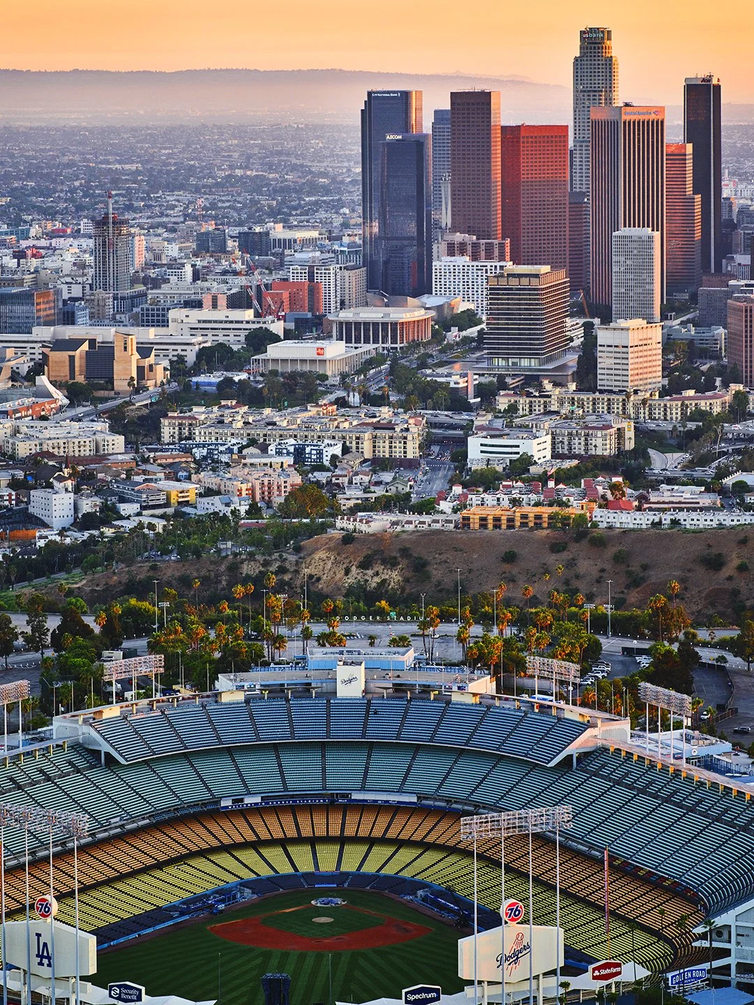 View of downtown Los Angeles with skyscrapers and the Dodger Stadium in the foreground at sunset.