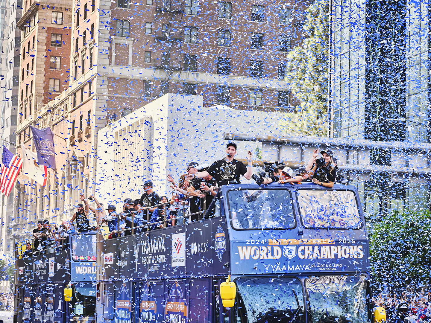 Celebrating sports team parade with players and fans on a double-decker bus, confetti falling, city buildings in background, and people waving and cheering.