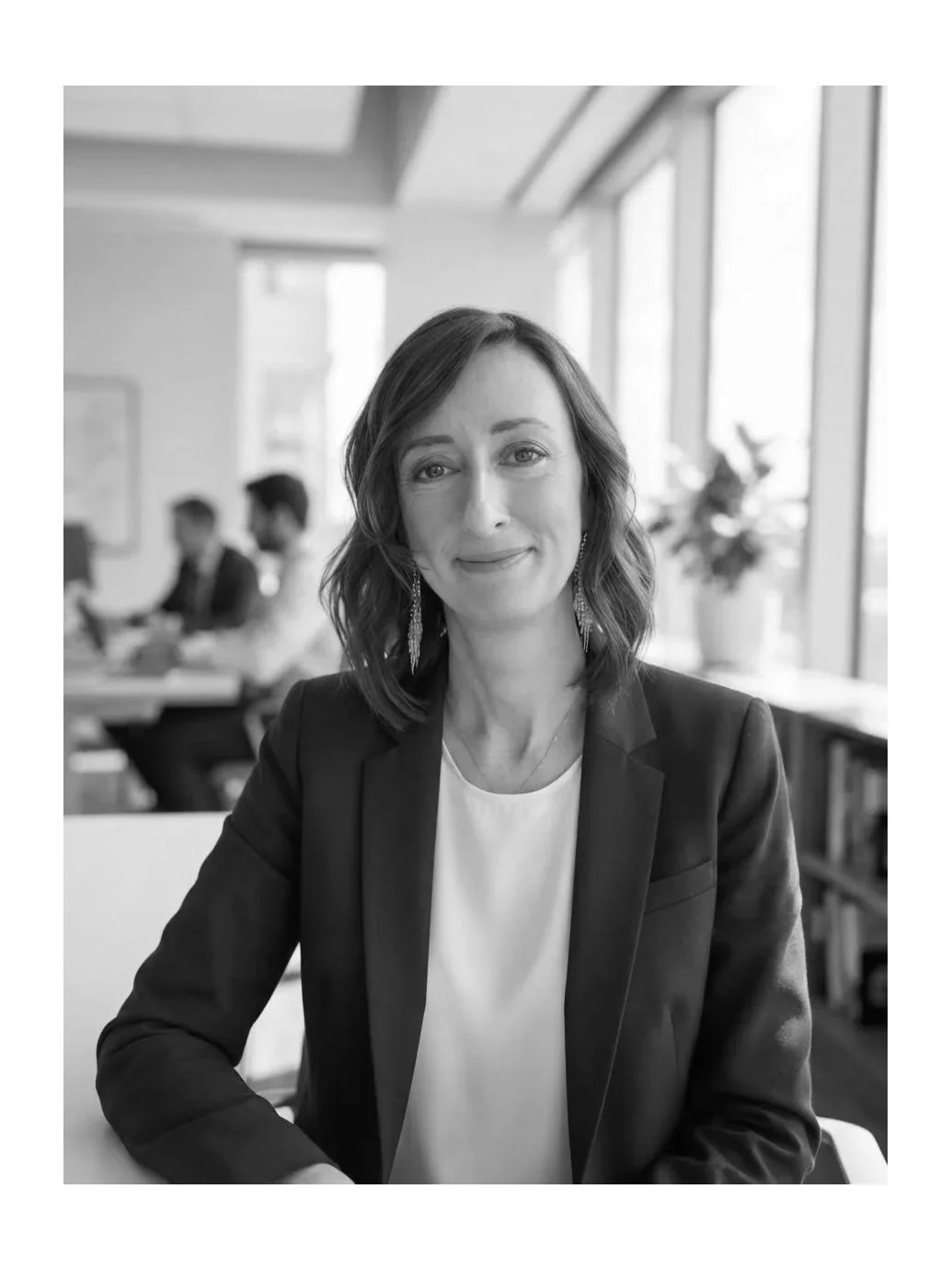 Black and white photo of a woman with shoulder-length hair, wearing a blazer and earrings, smiling at the camera, in an office setting with large windows and bookshelves in the background.